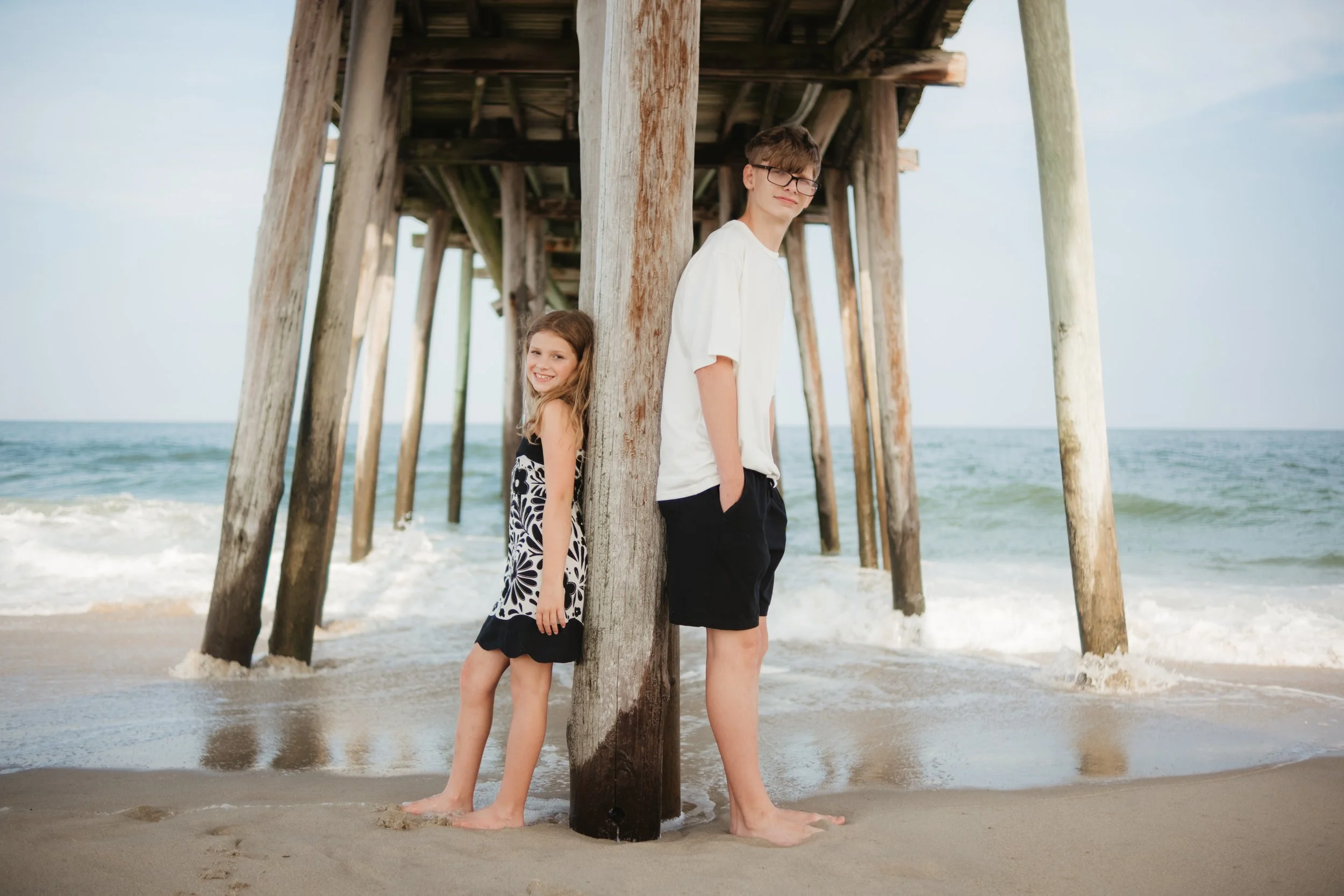 A boy and girl stand back-to-back under a wooden pier on the beach, with the ocean in the background. The girl is smiling, wearing a black and white dress, and the boy is looking at the camera, wearing glasses, a white t-shirt, and black shorts.