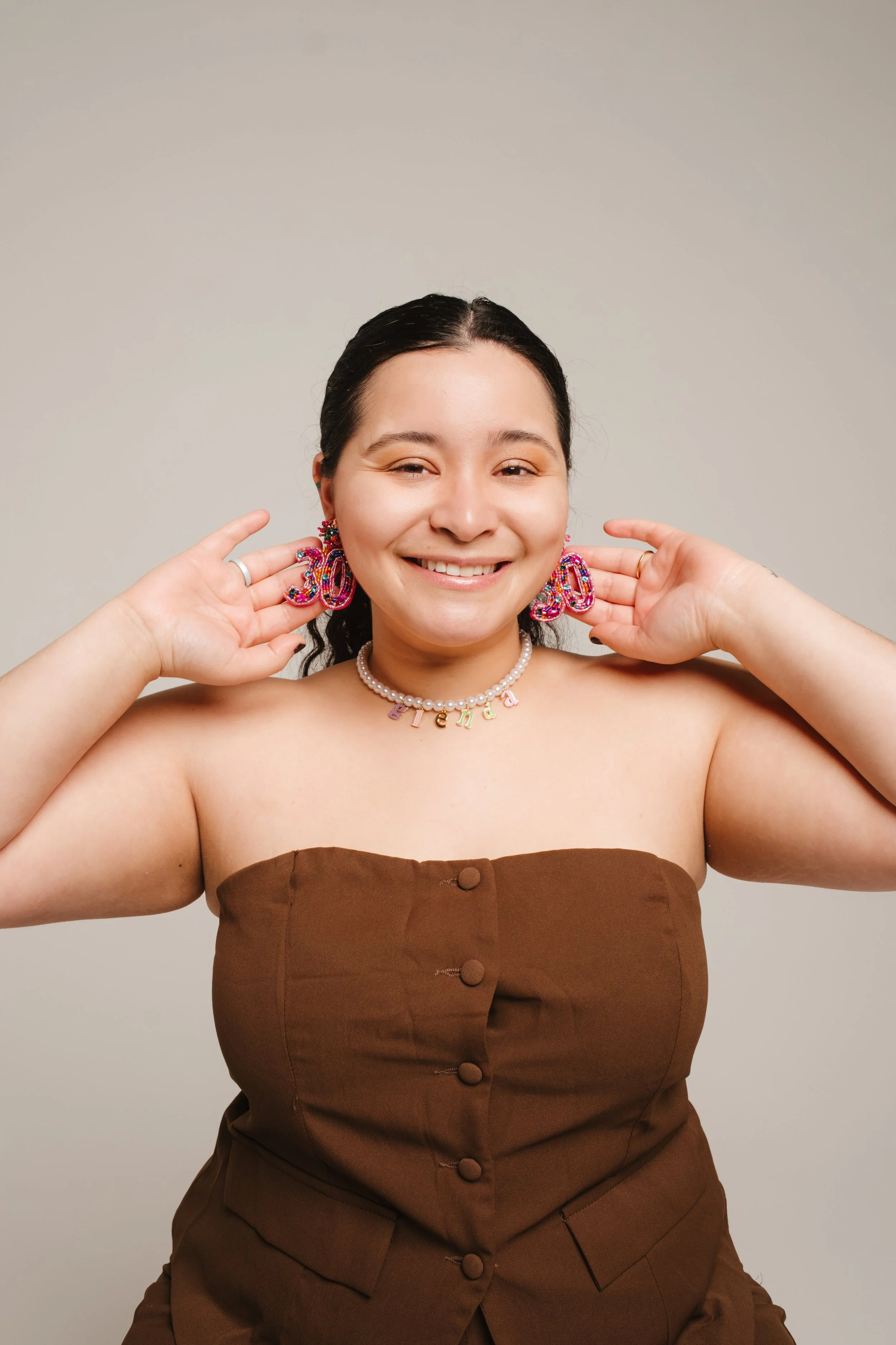A young woman smiling, wearing a strapless brown dress, colorful beaded earrings, a pearl necklace with "friend" spelled out in beads, and holding her hands near her ears against a plain neutral background.