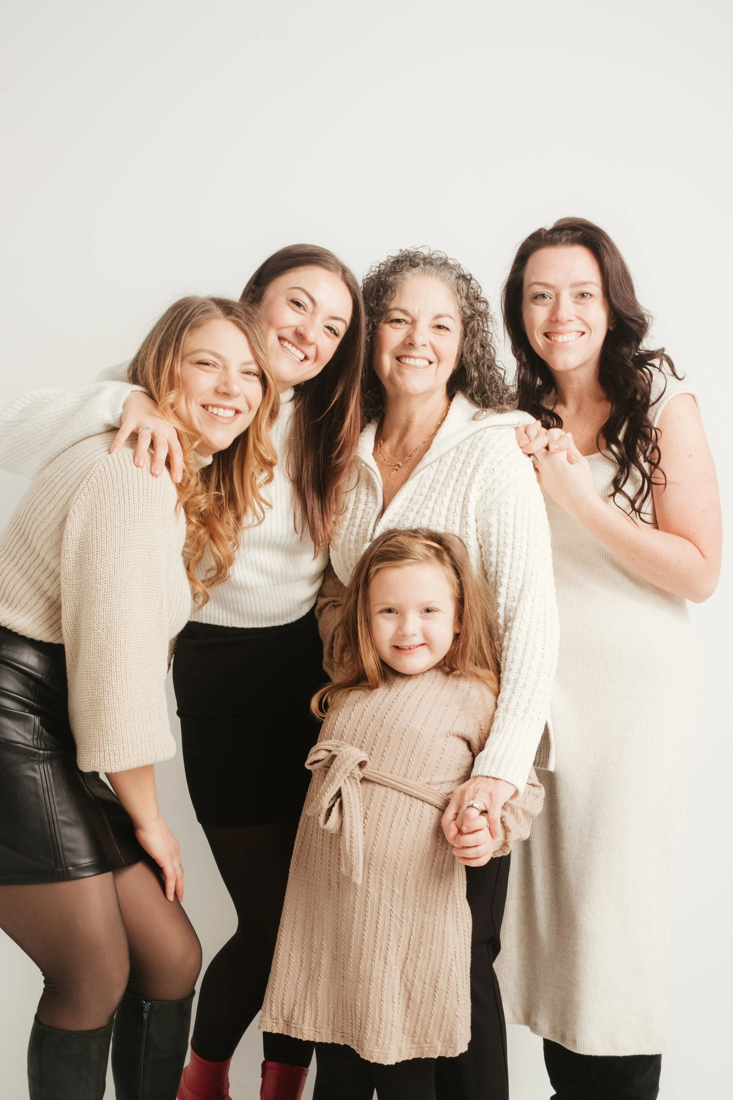 Group of six women and a girl smiling and hugging together in a white studio.