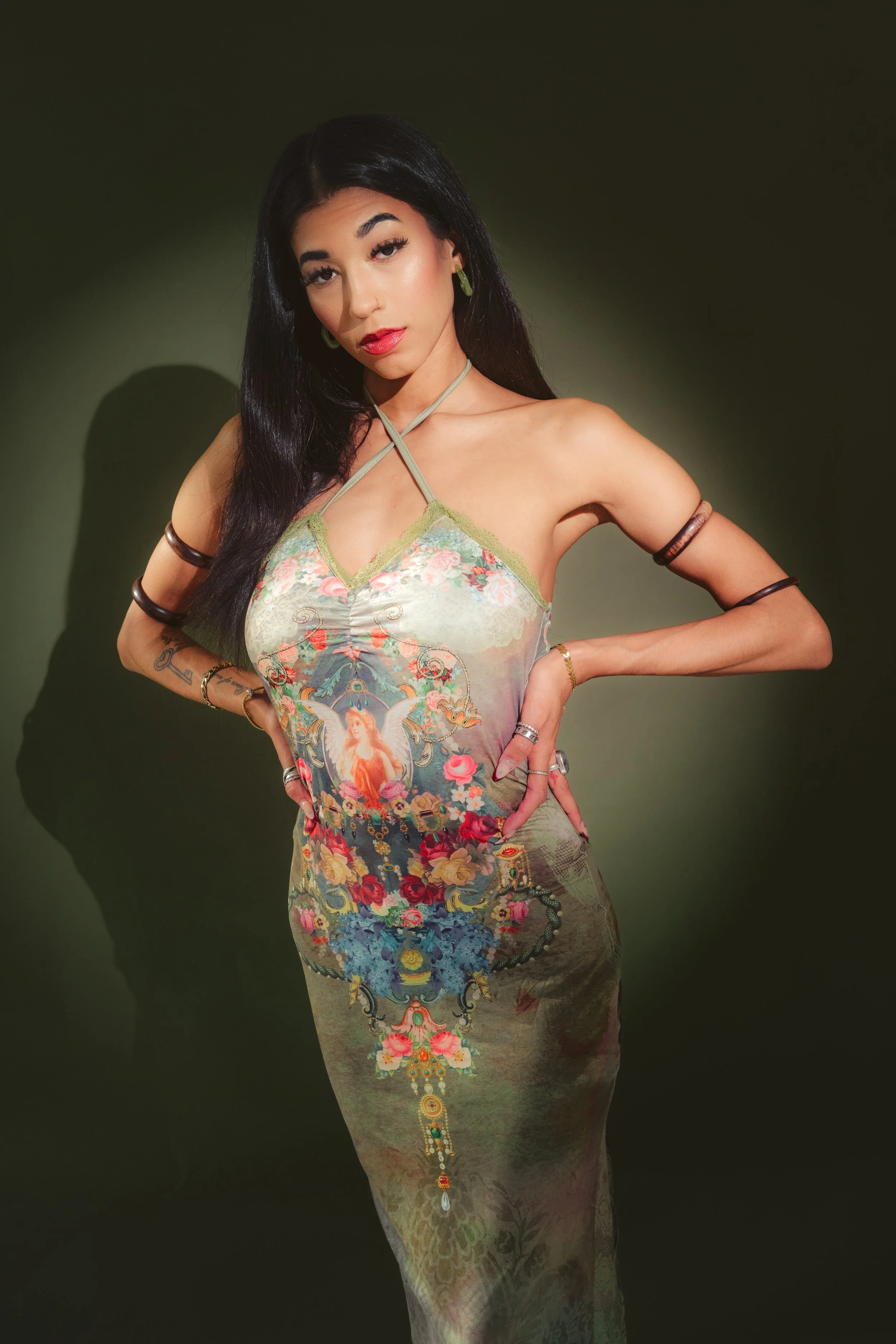 A woman with long black hair in a floral dress with symbols and images, posing with hands on hips against a dark green background.