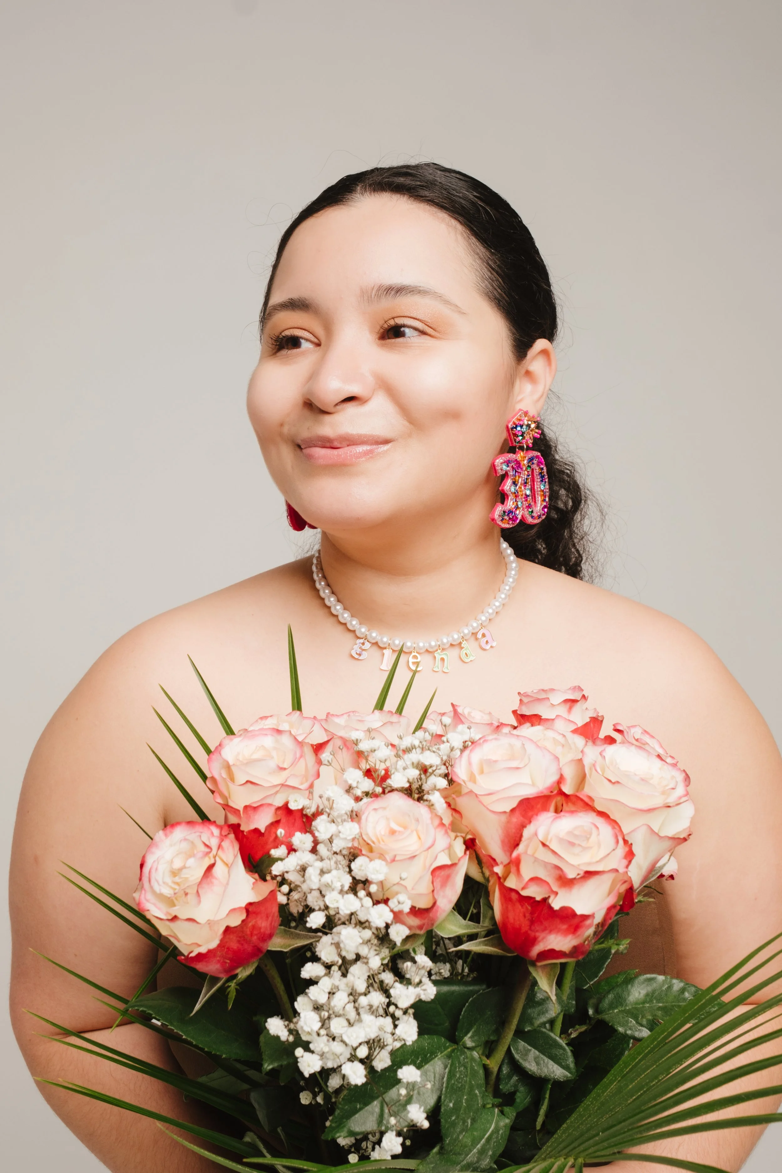 A woman holding a bouquet of pink and white roses with greenery, wearing colorful earrings, a pearl necklace, and a 'fiesta' name necklace, smiling and looking to her right against a plain background.