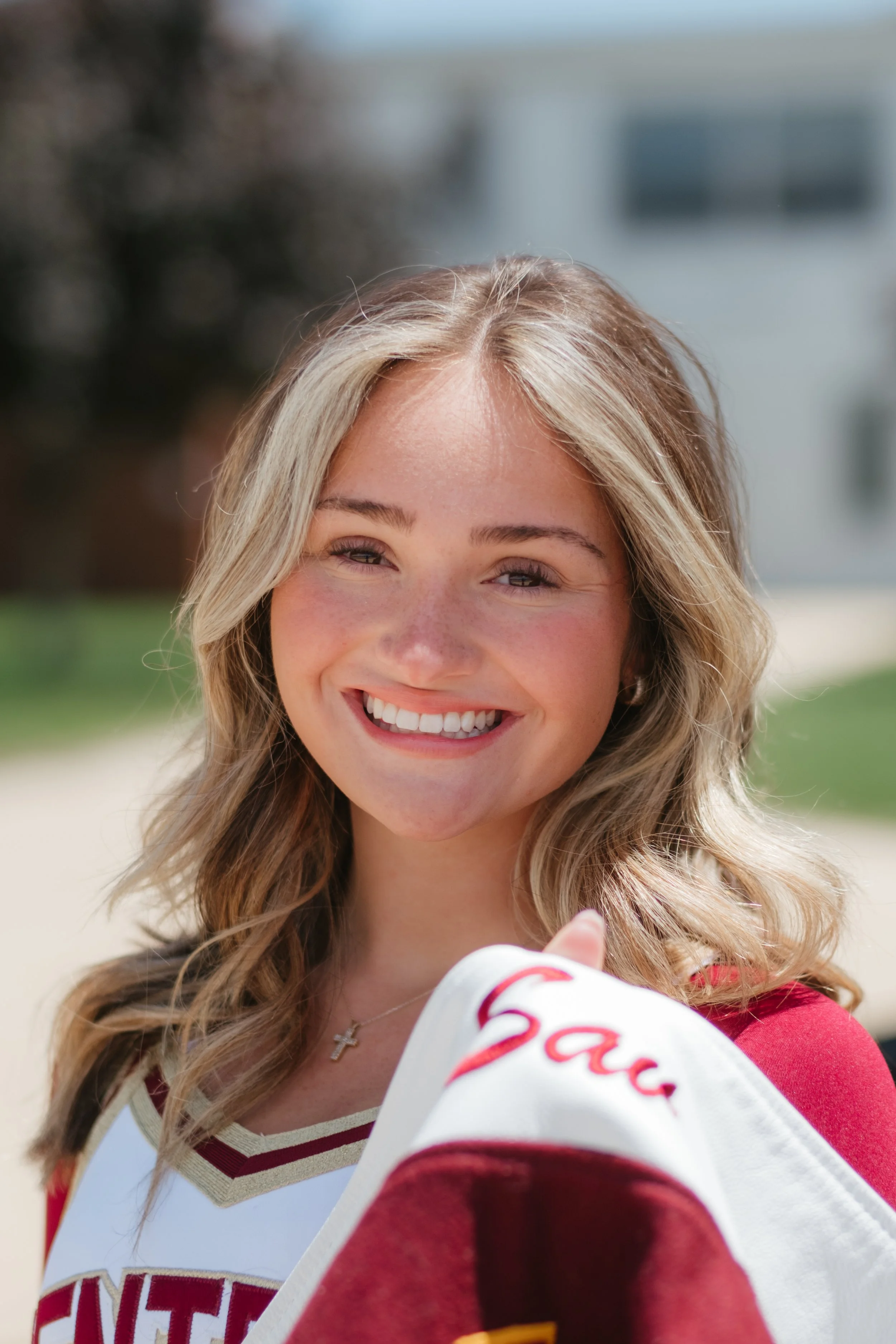 A young woman with blonde, wavy hair smiling outdoors wearing a red and white sports jersey.