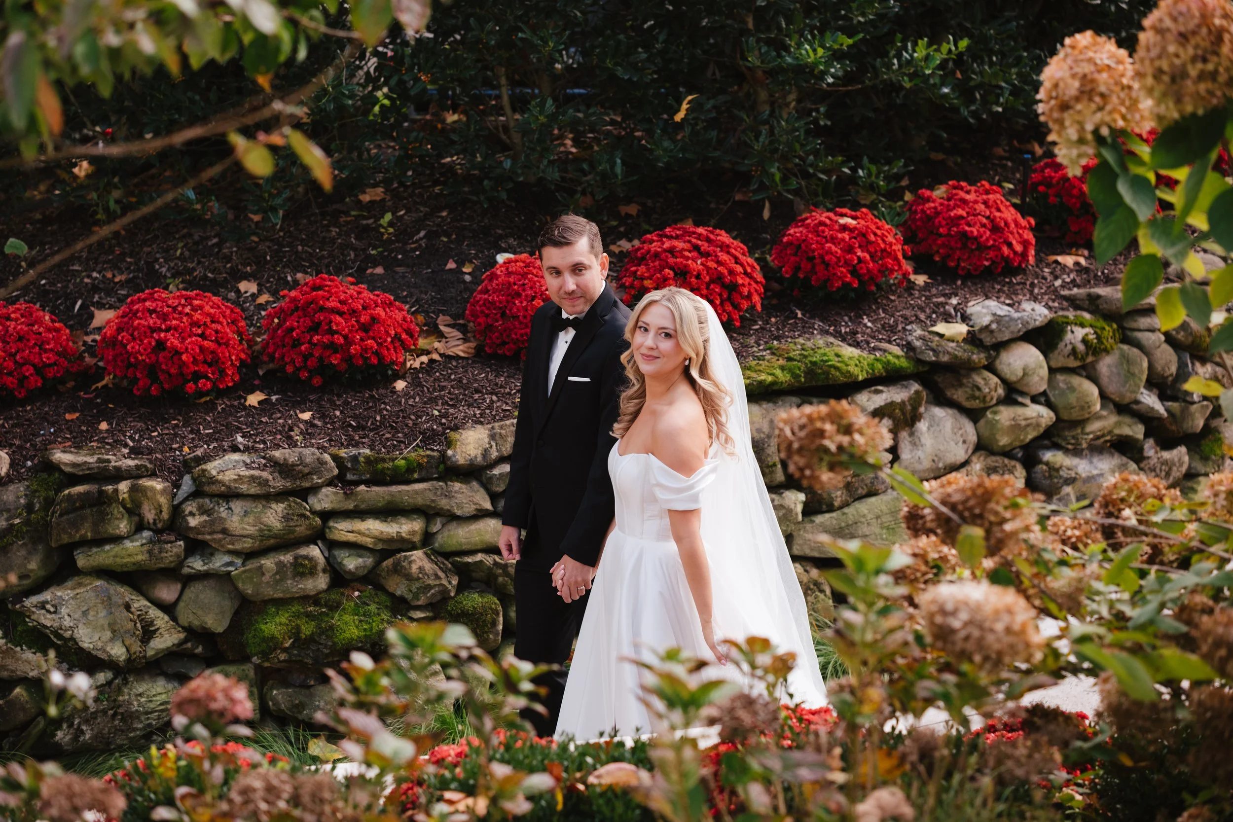 A bride and groom holding hands in a garden, with vibrant red and brown hydrangeas and rocks in the background.