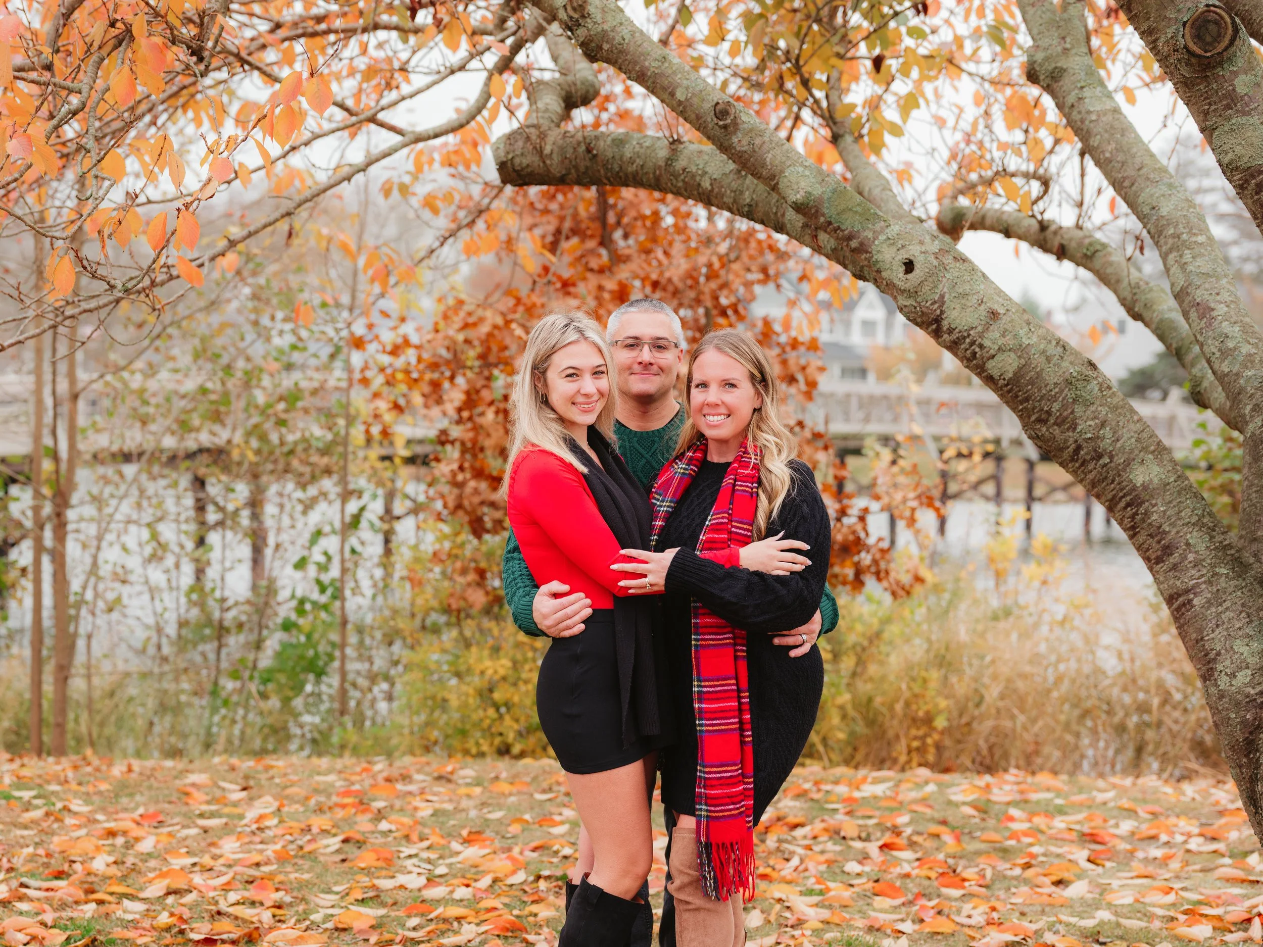 A family of three, two women and a man, standing outside in autumn surrounded by orange and brown leaves, under a large tree with twisting branches, smiling at the camera.