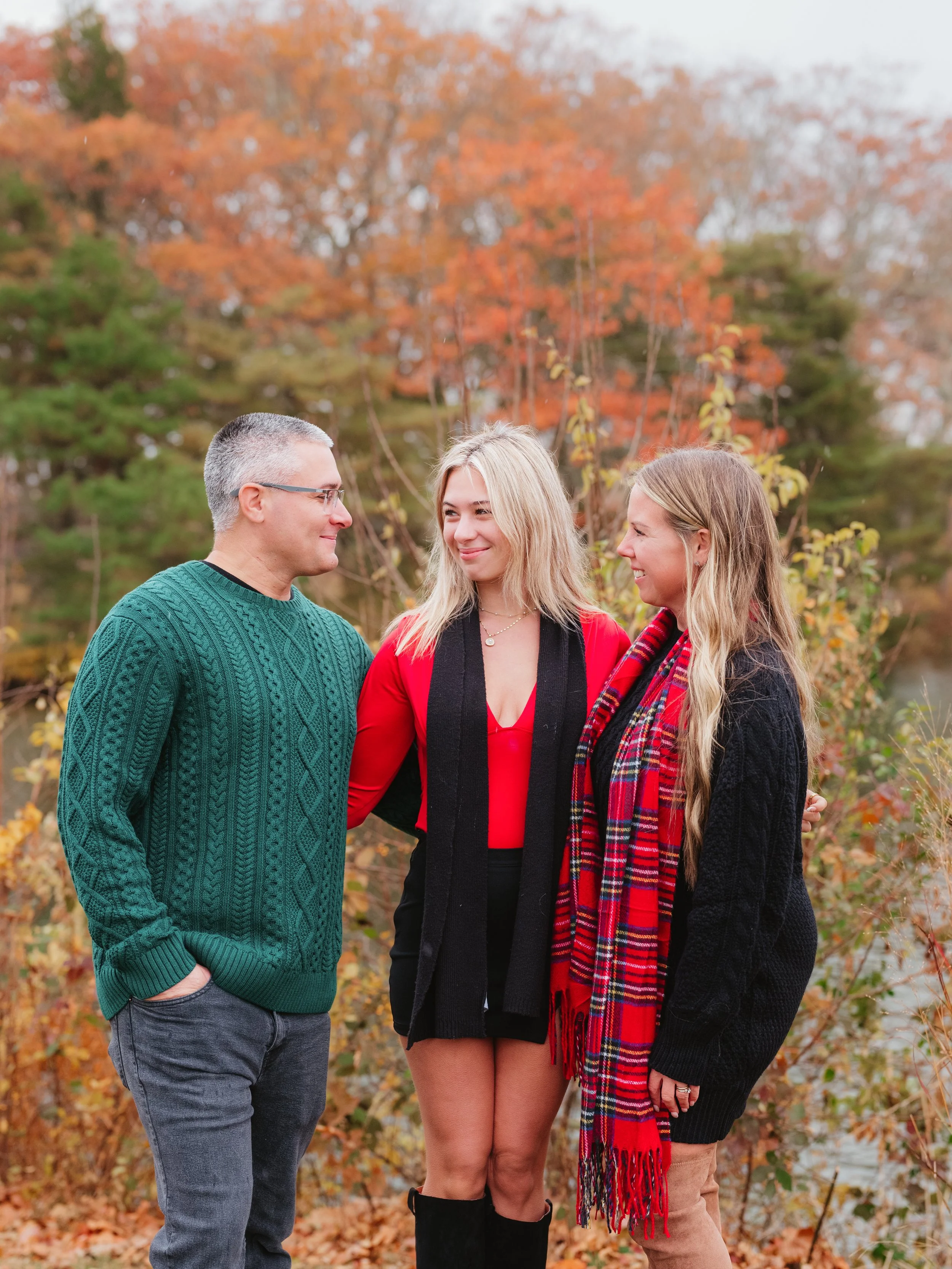 Three people standing outdoors in fall foliage, smiling and looking at each other.