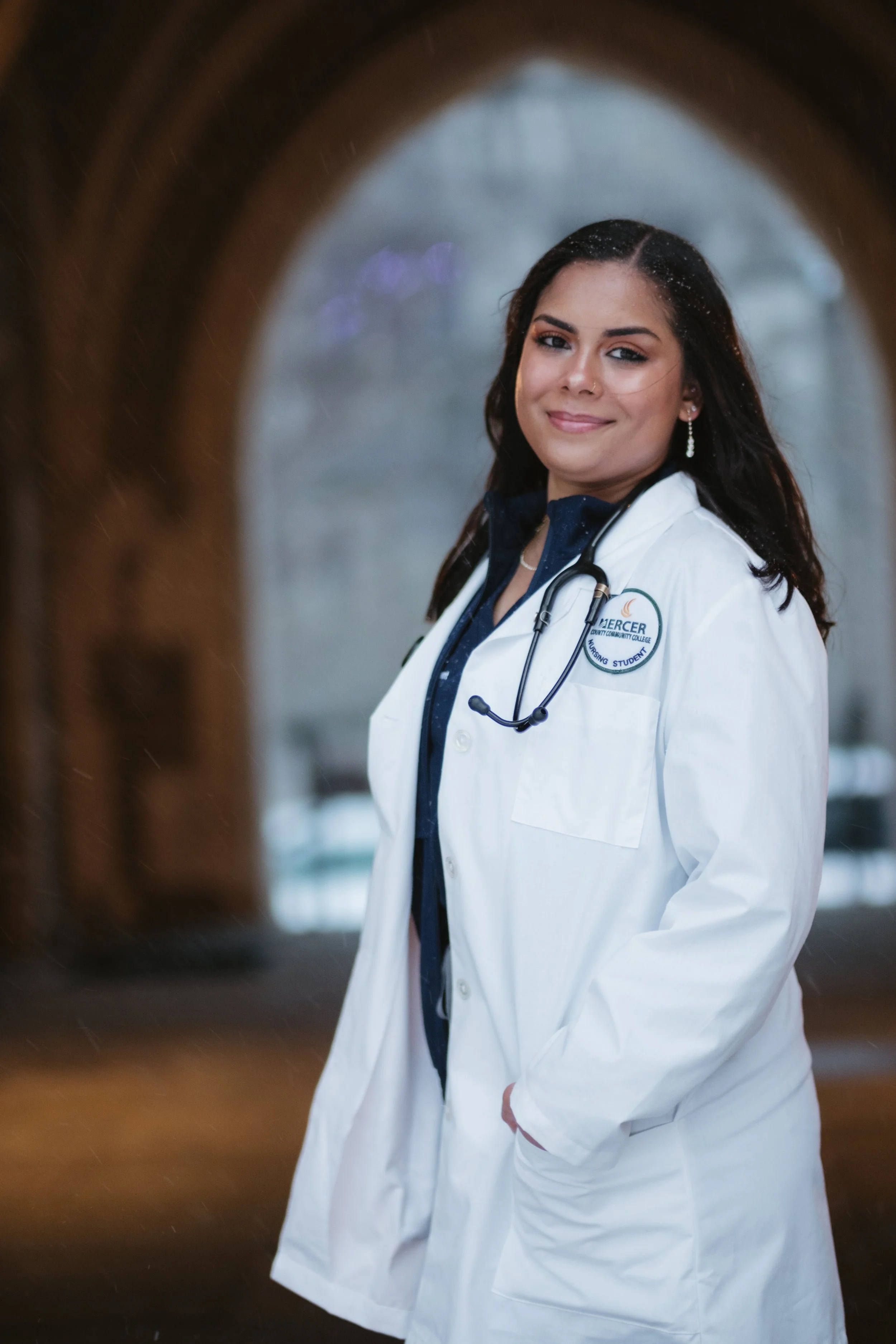 A smiling female doctor with dark hair, wearing a white lab coat and stethoscope, standing in front of an arched window with rain outside.