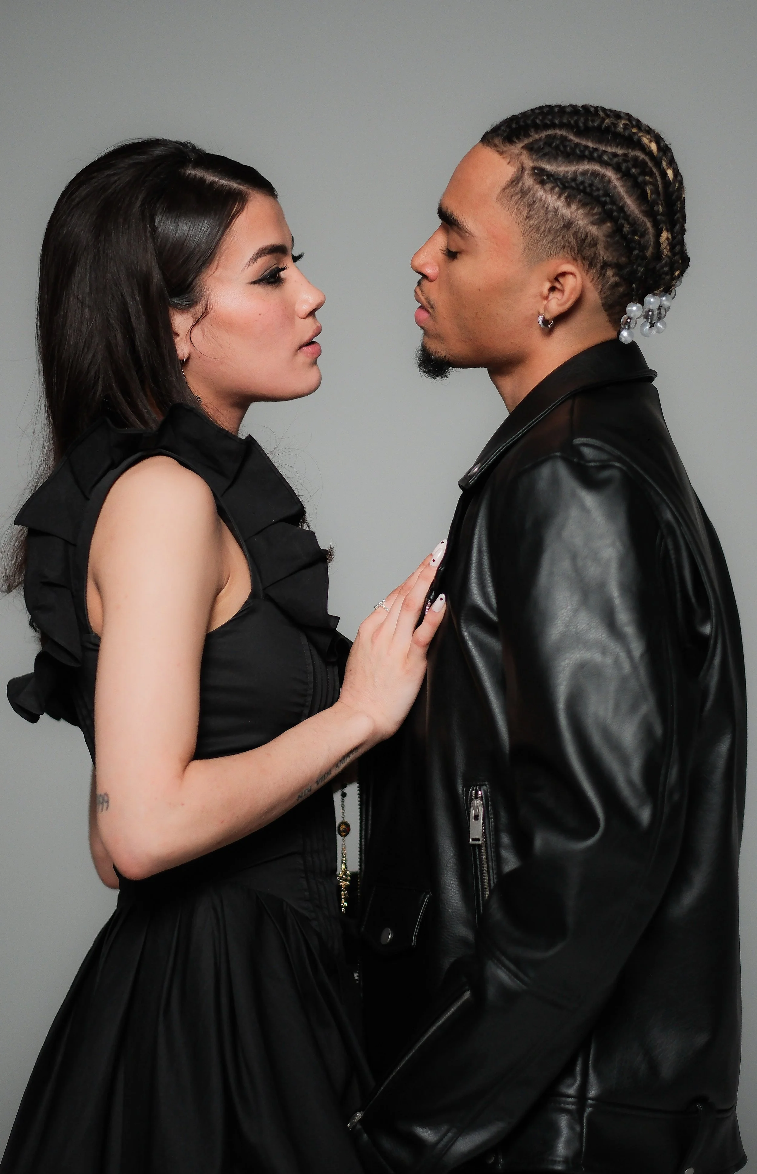 A woman and a man face each other closely, the woman touching the man's chest, both with serious expressions, against a plain gray background.