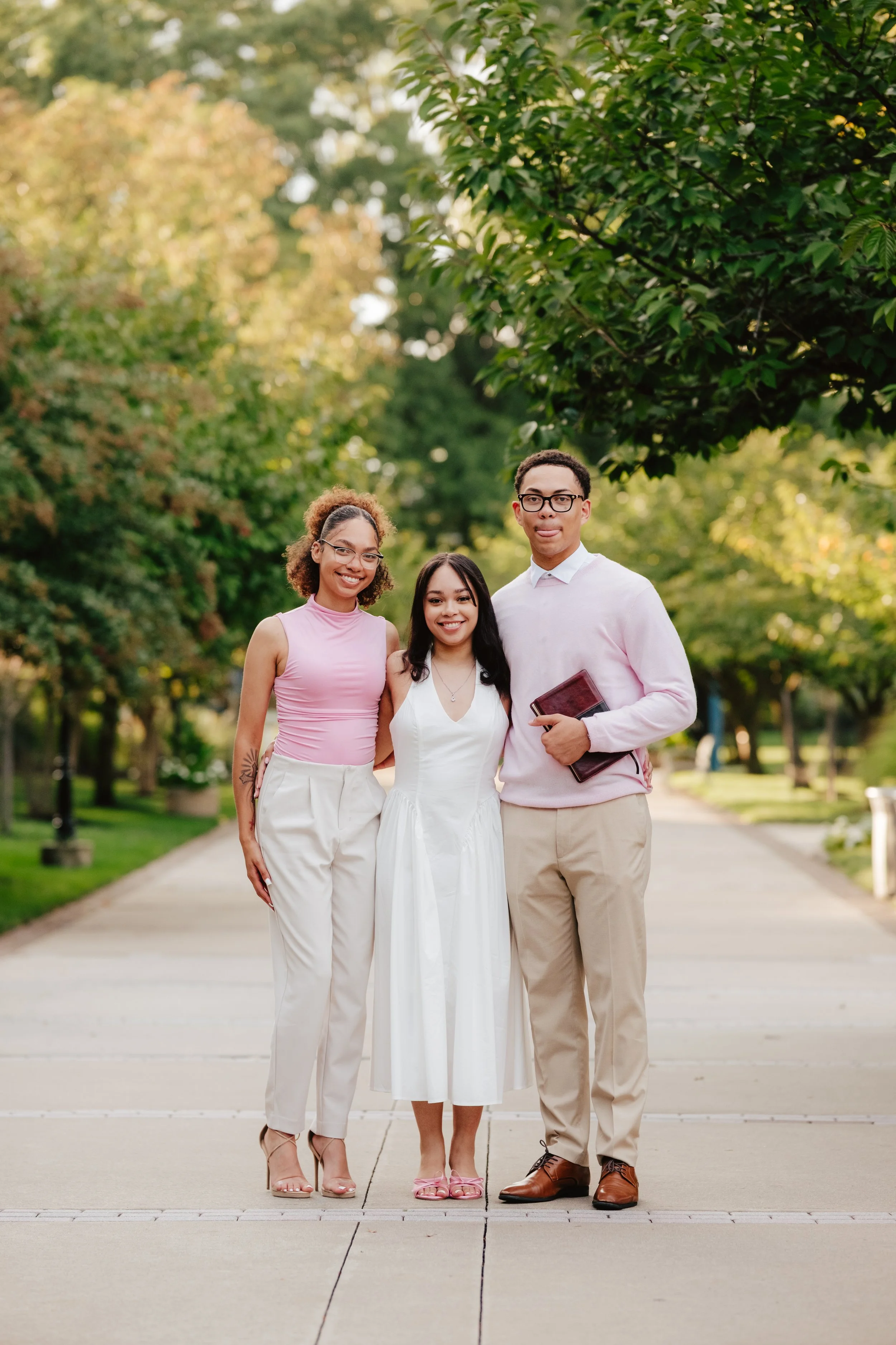 Three young adults standing arm in arm on a sidewalk in a park with green trees, smiling at the camera.