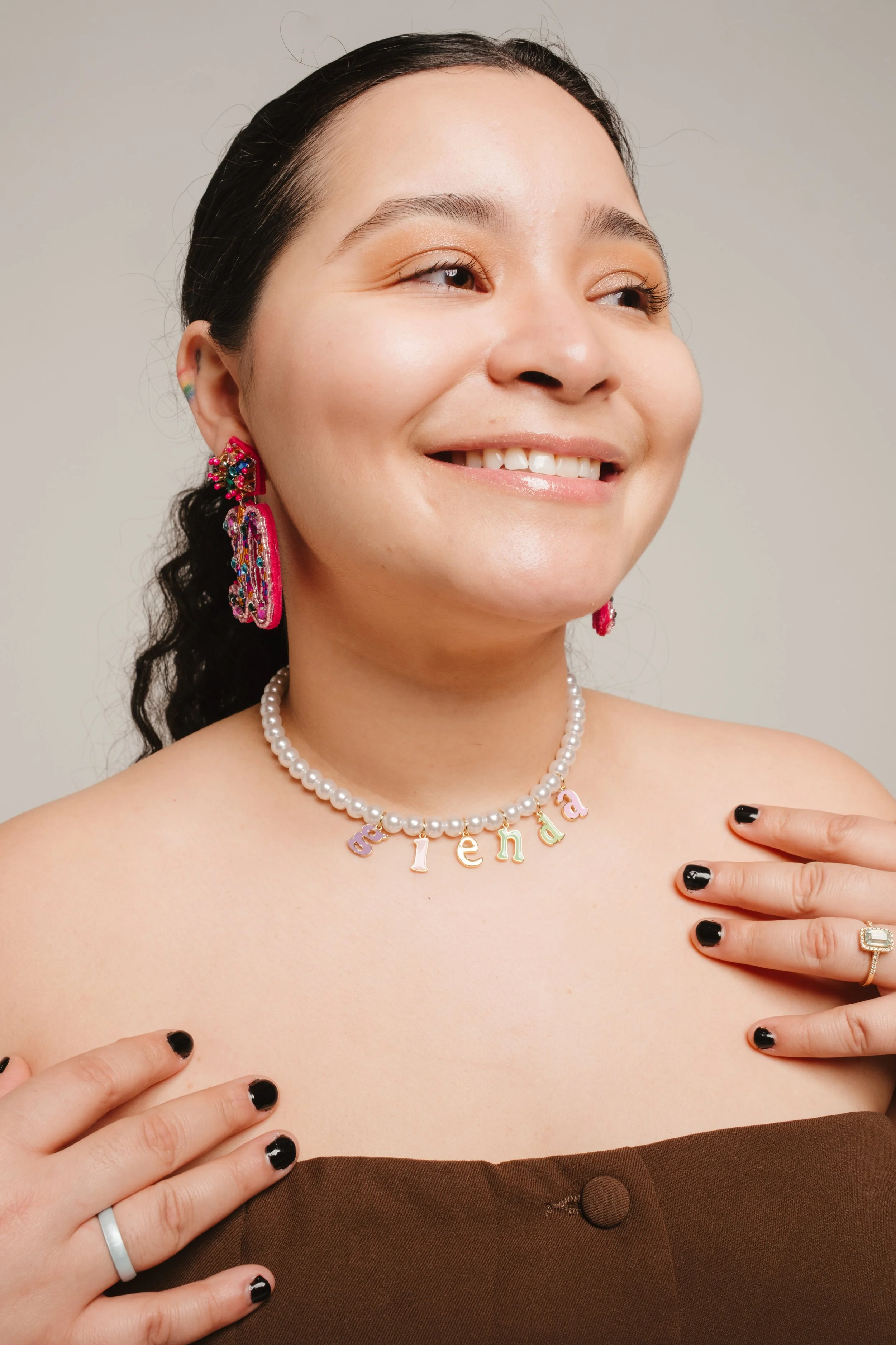 A woman with black hair tied back, wearing colorful earrings, a pearl necklace spelling 'arenda', a brown strapless top, and rings on her fingers, smiling and touching her shoulders.