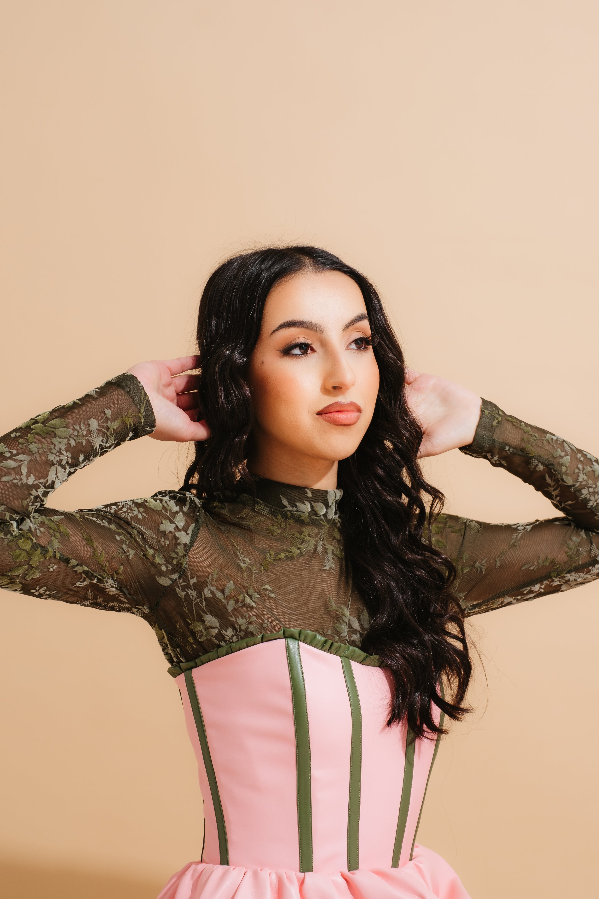 Woman with long dark wavy hair posing with hands near her head wearing a sheer green floral top and a pink corset-style dress with green vertical stripes, standing against a beige background.