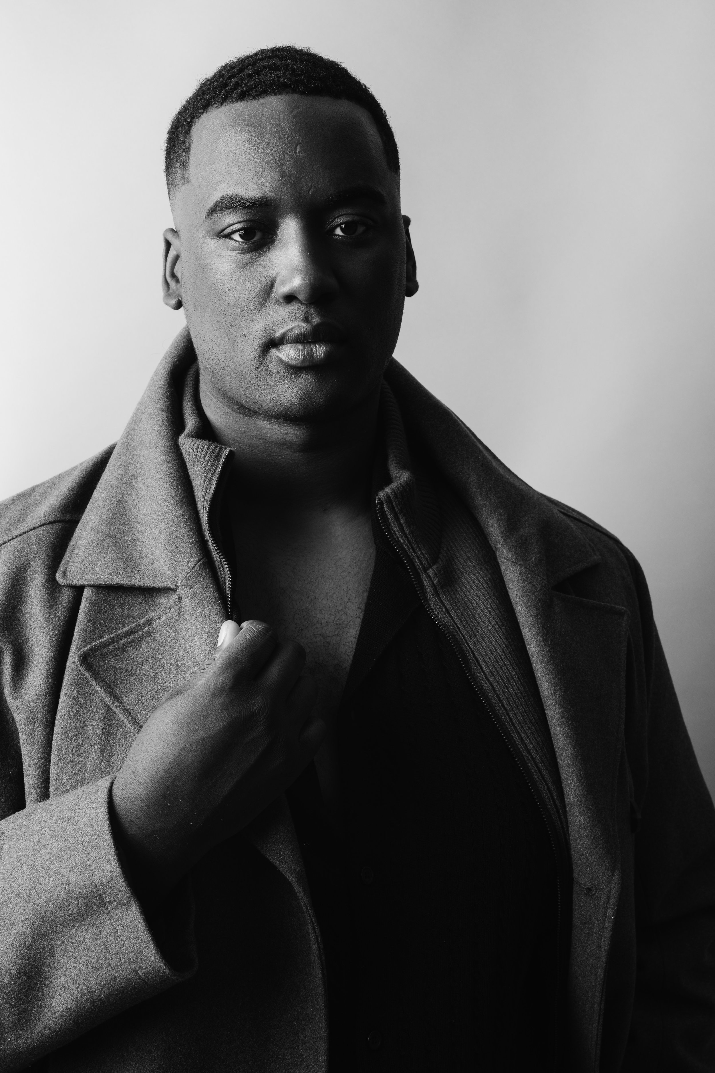 Black and white portrait of a young man with short hair, wearing a dark jacket and a zip-up sweater, holding the collar of his coat with his left hand, looking directly at the camera against a plain background.