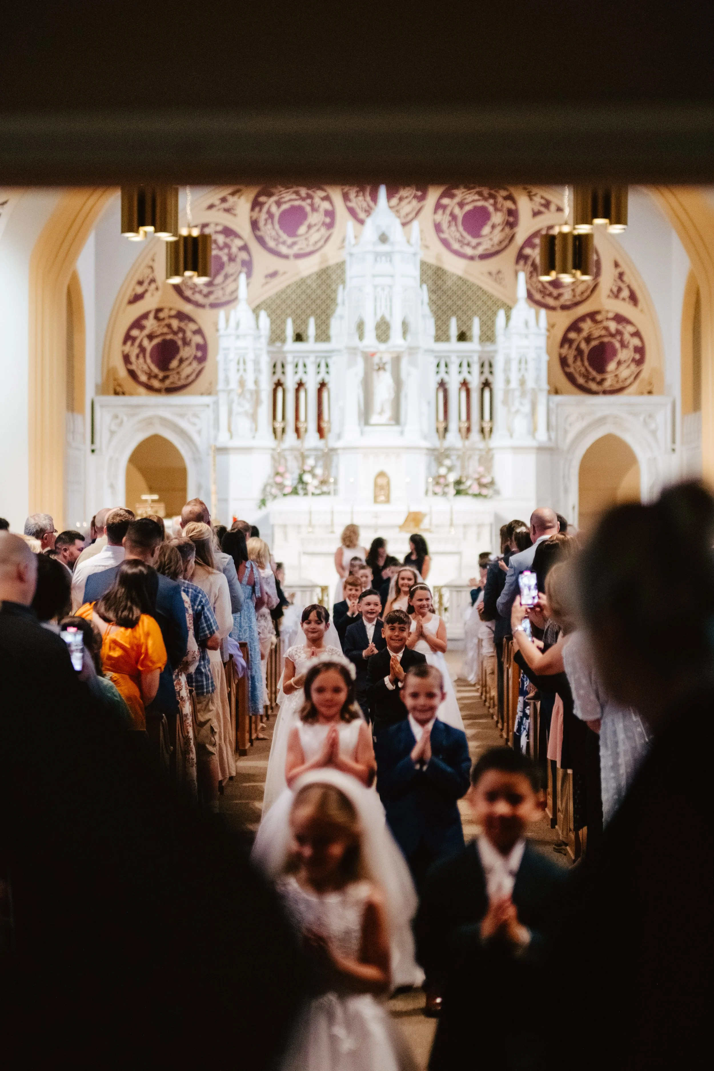 Children dressed in formal attire walking down the aisle of a church, surrounded by adults taking photos, with an ornate altar in the background.