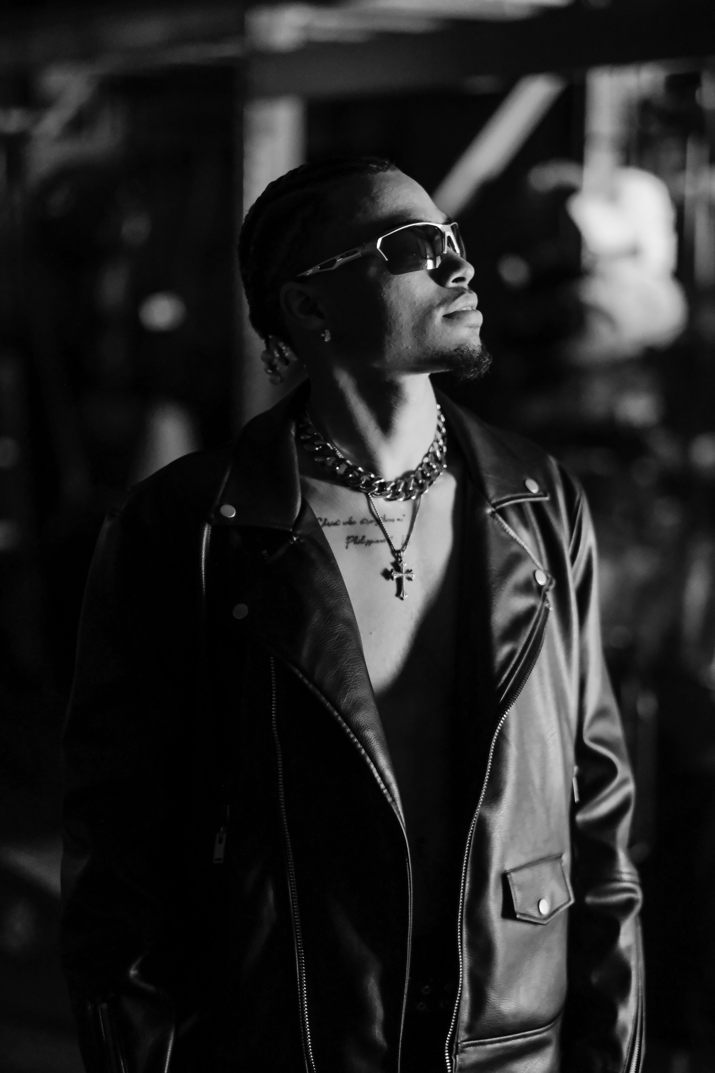 A man wearing sunglasses, a leather jacket, and chunky jewelry, including a chain necklace and a cross pendant, standing outdoors at night with a blurred background.