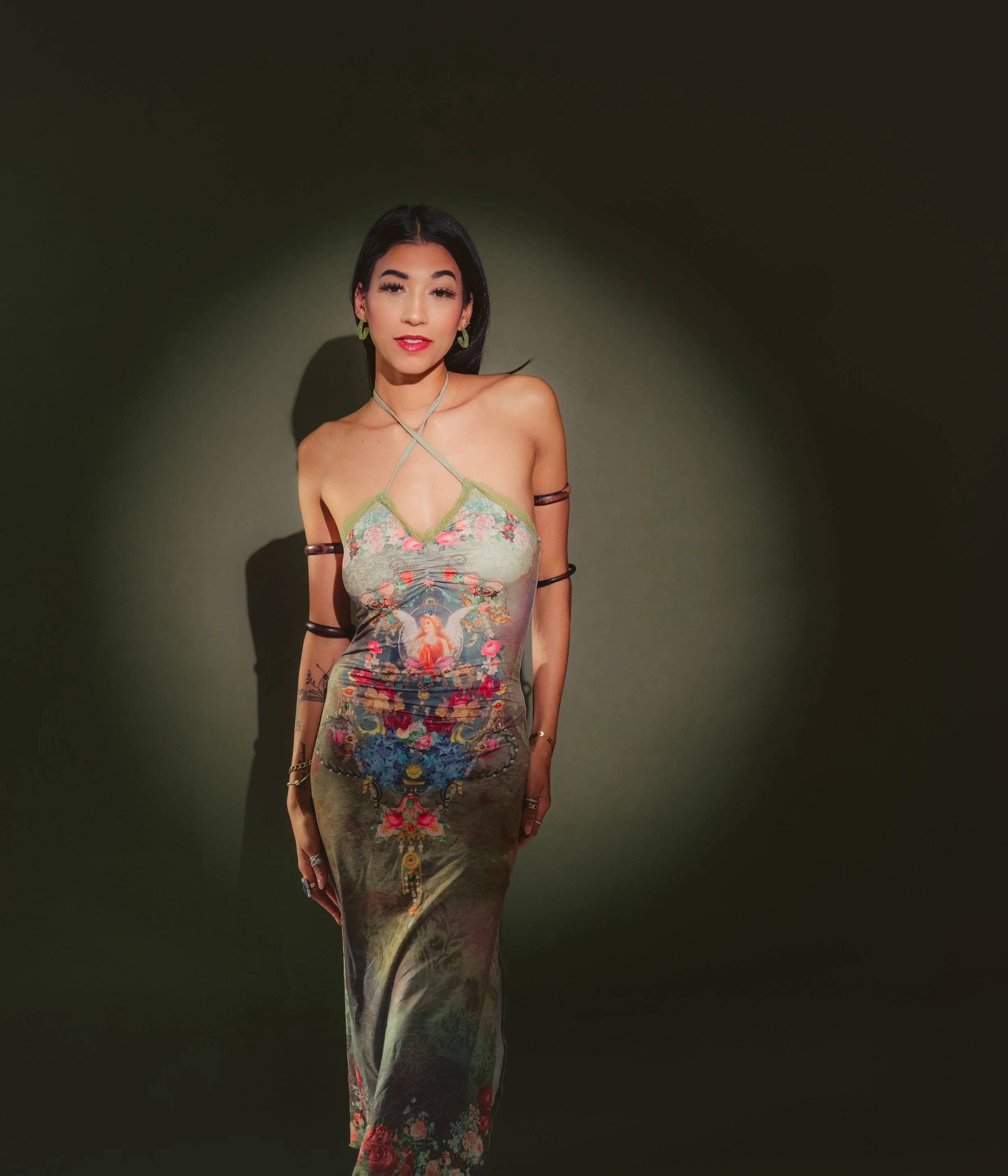 A woman standing against a dark background, wearing a colorful, patterned dress with thin straps and an artistic design.