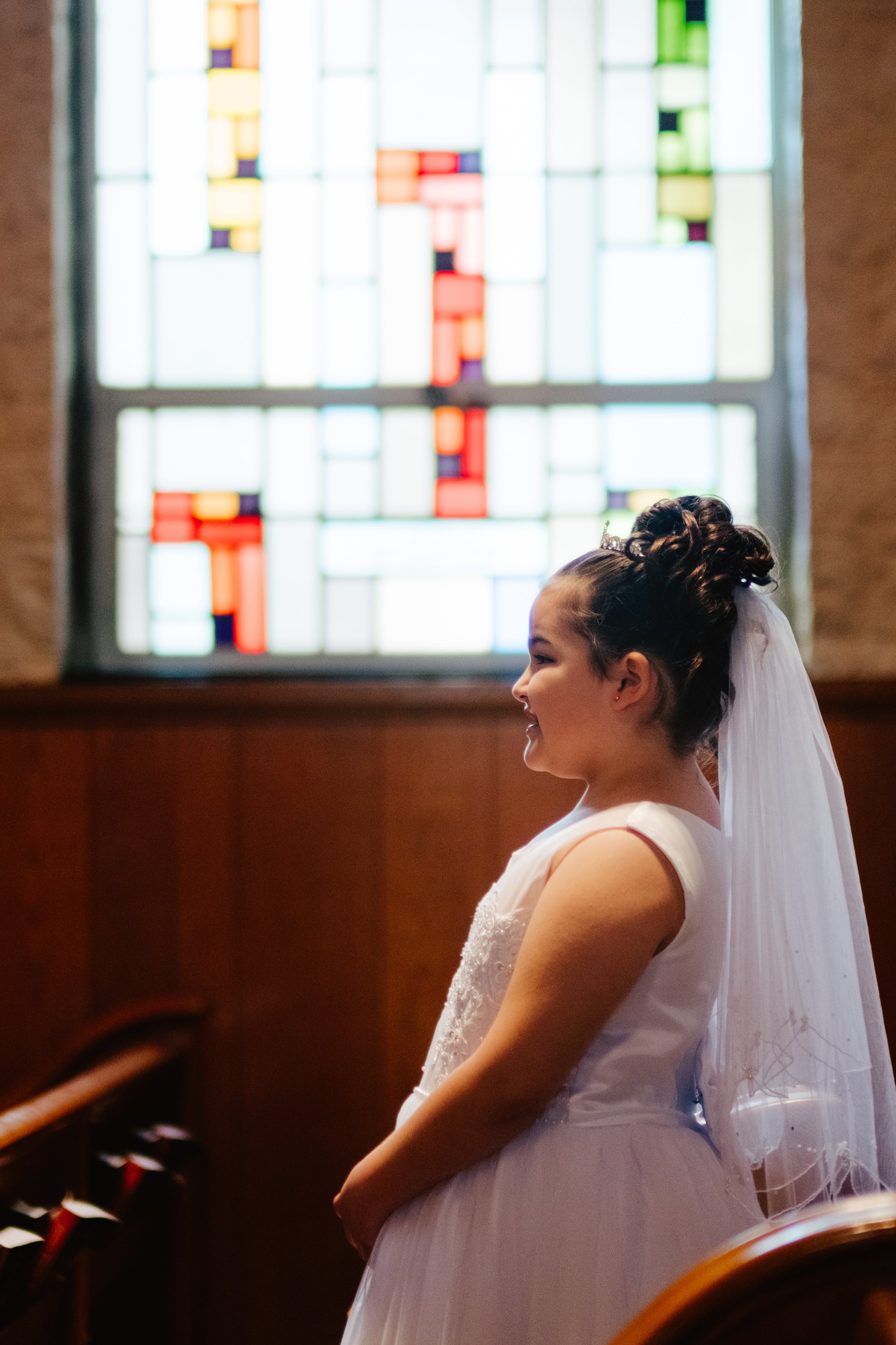 A girl dressed as a bride with a white dress and veil, standing inside a church with a stained glass window in the background.