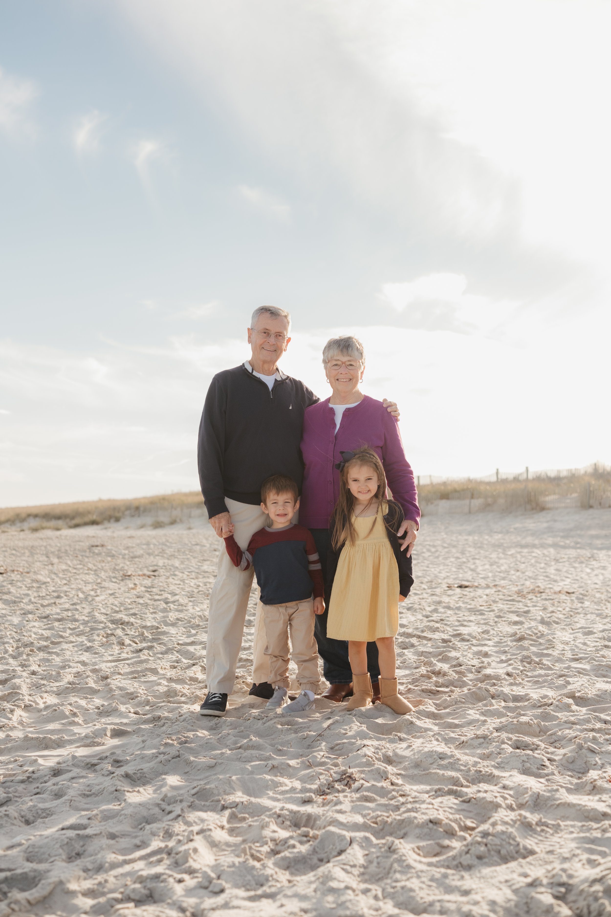 A happy multi-generational family of four standing on the beach, smiling at the camera, with a cloudy sky in the background.