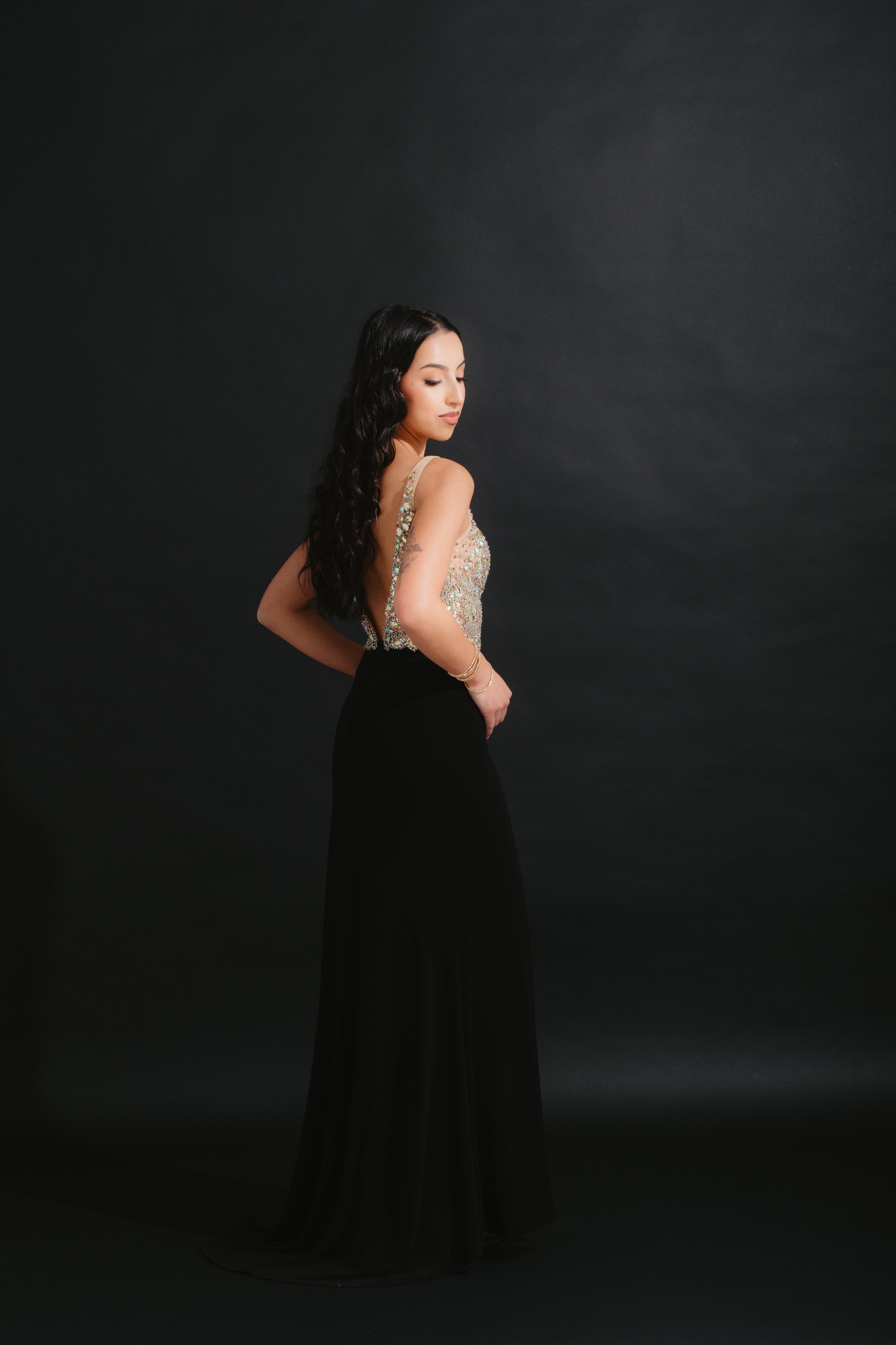 A young woman with long dark hair in an elegant dress poses against a dark background.