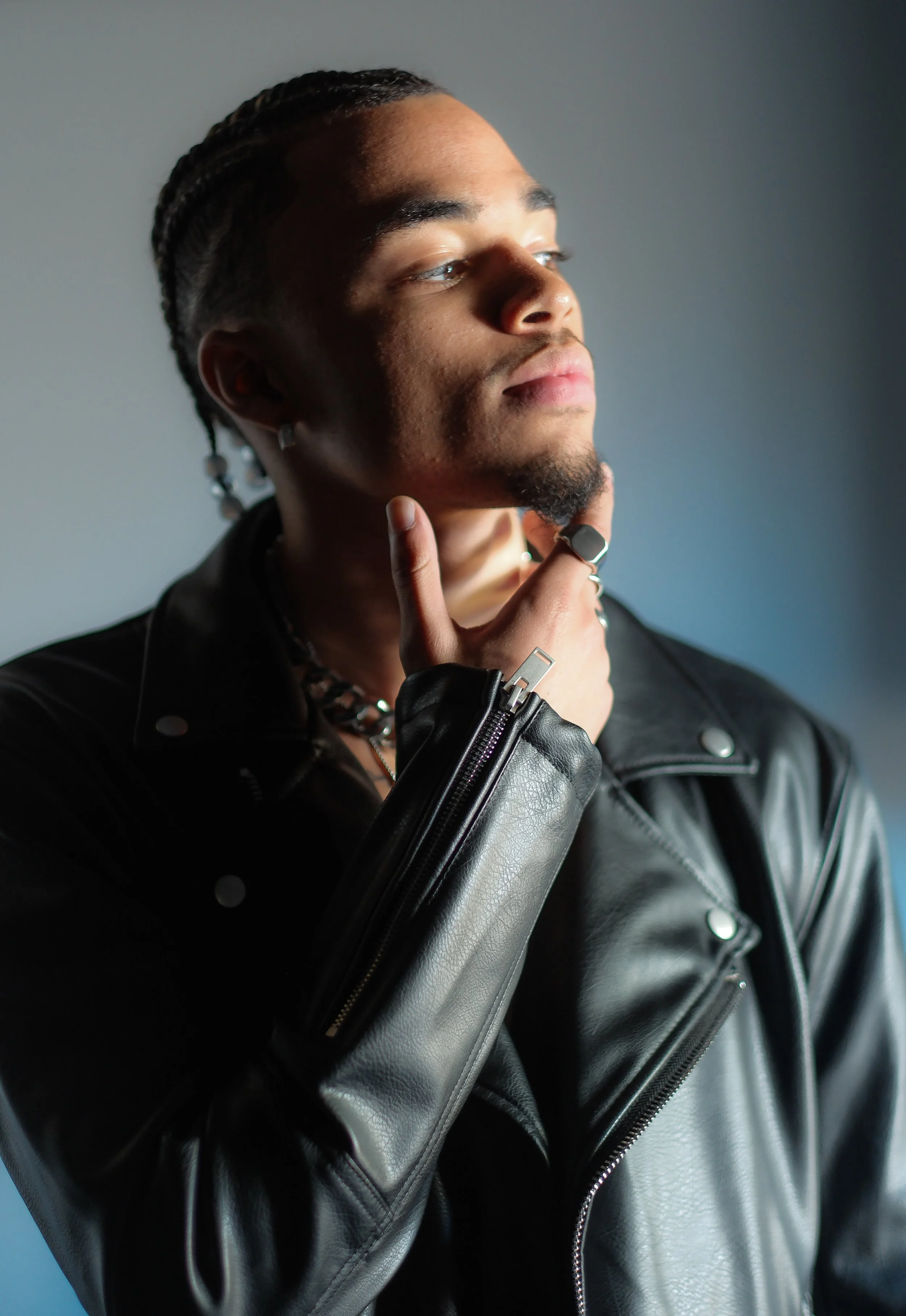 A young man with braided hair and earrings, wearing a black leather jacket, is posing thoughtfully with his hand on his chin against a neutral background.