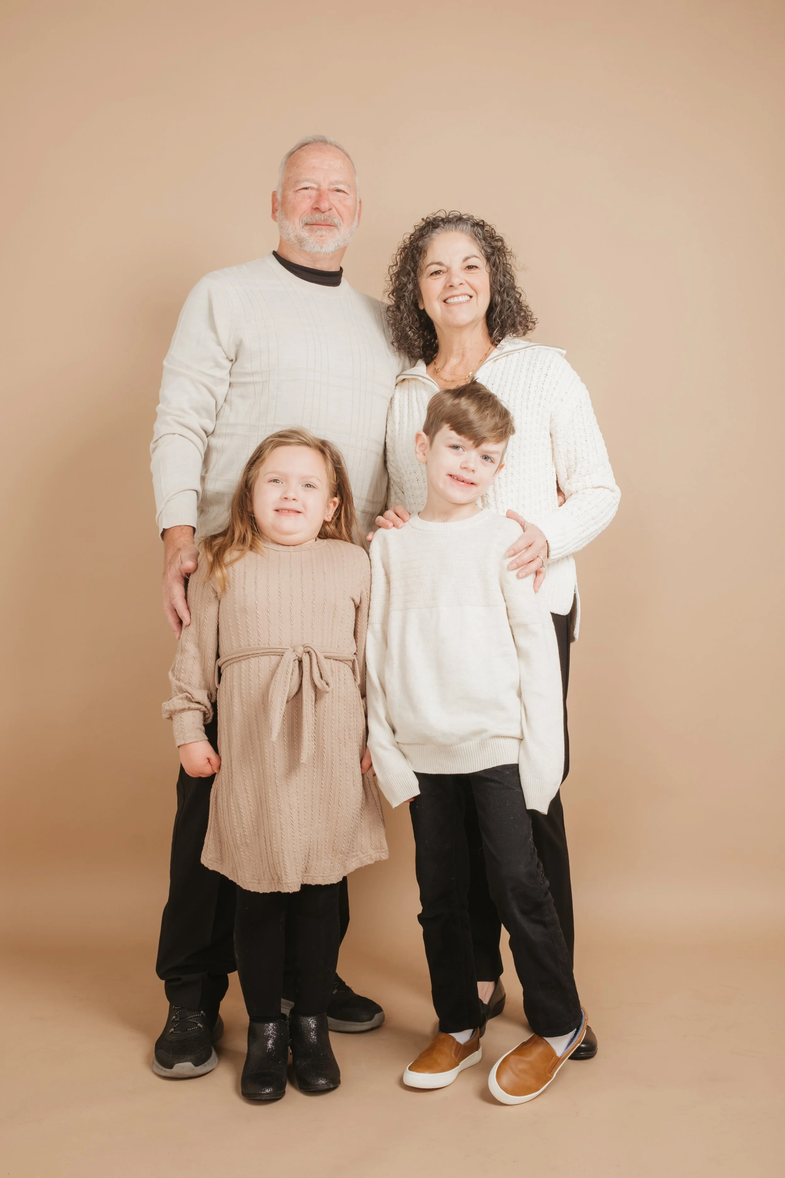 A family of five standing together against a beige background, smiling at the camera. The group includes an older man and woman, and a young boy and girl.