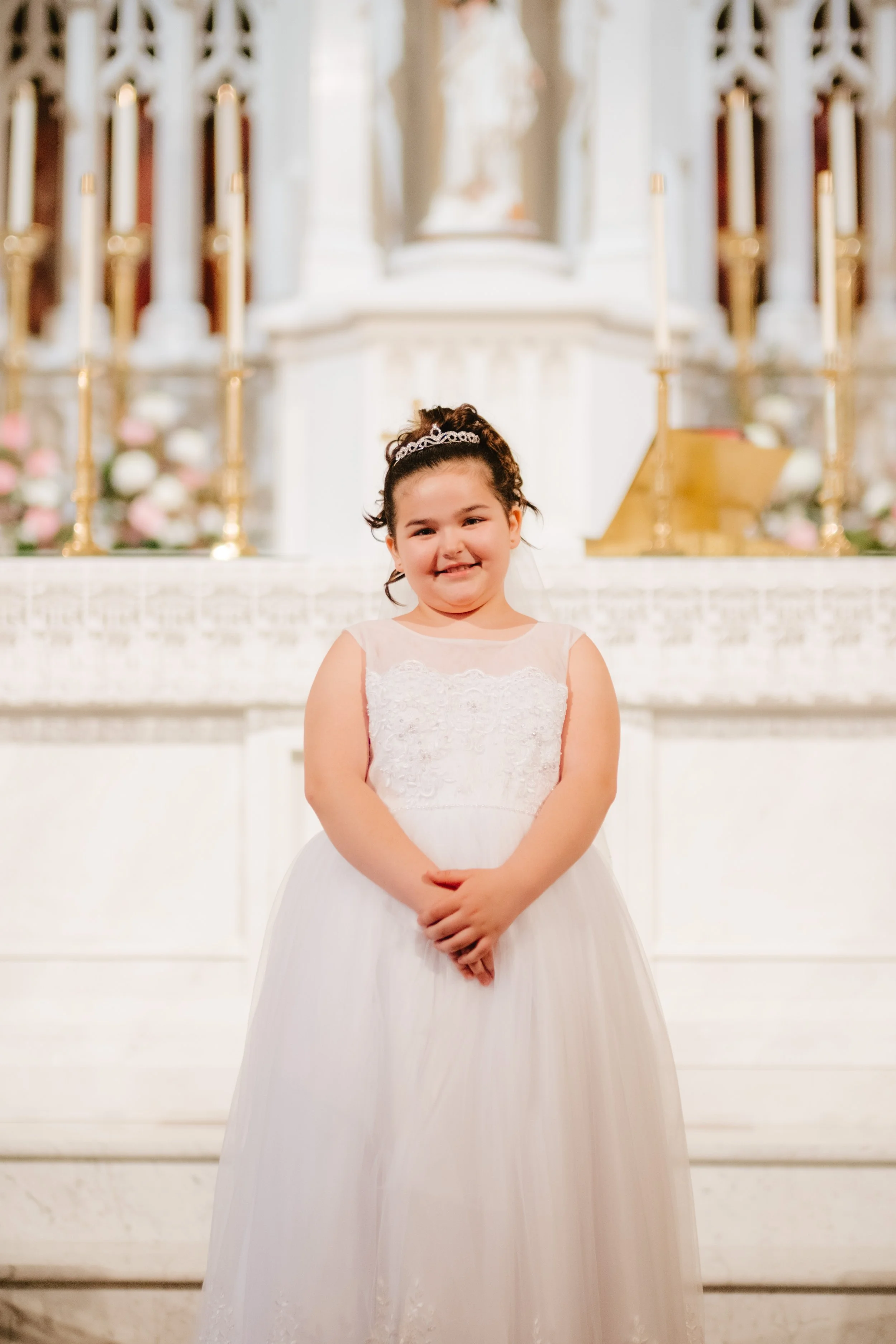 A young girl in a white dress with lace, standing in a church or cathedral with an altar, candles, and floral arrangements in the background.