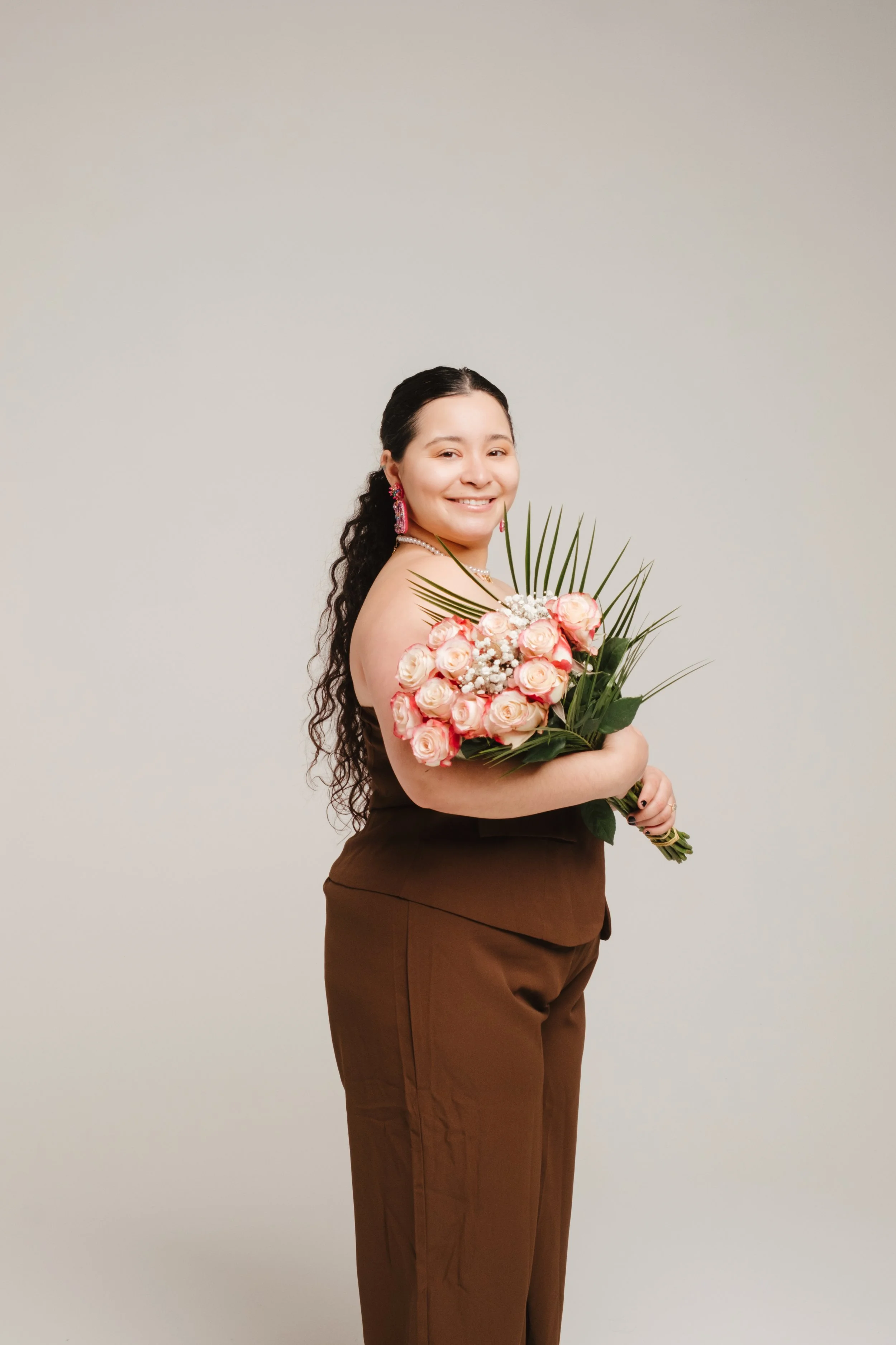 Woman with long dark curly hair smiling, wearing brown pants, holding a bouquet of pink roses and greenery, standing against a plain light-colored background.