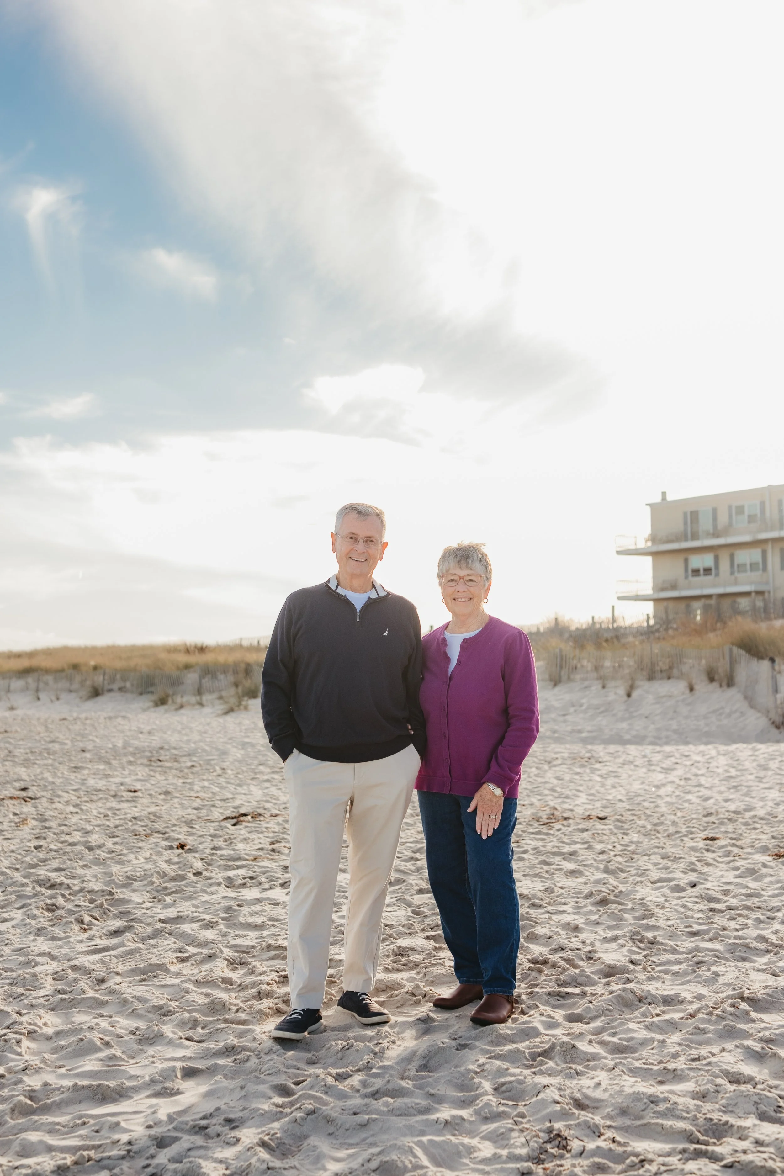 An older man and woman standing on a sandy beach, smiling at the camera, with a house on the dunes in the background under a cloudy sky.