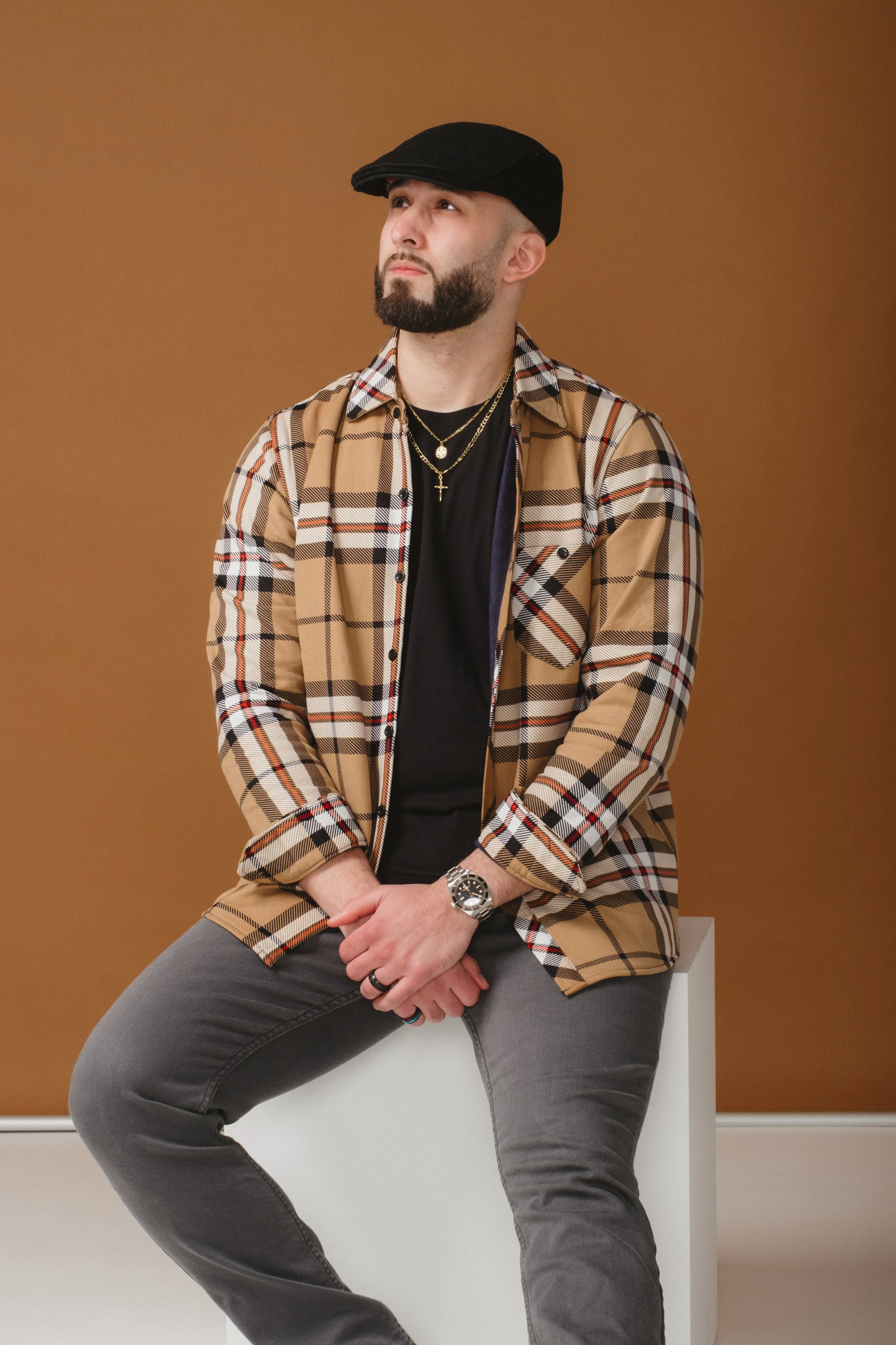 A man sitting against a brown background, wearing a black cap, checkered shirt, black t-shirt, gray jeans, and jewelry, looking to his left.