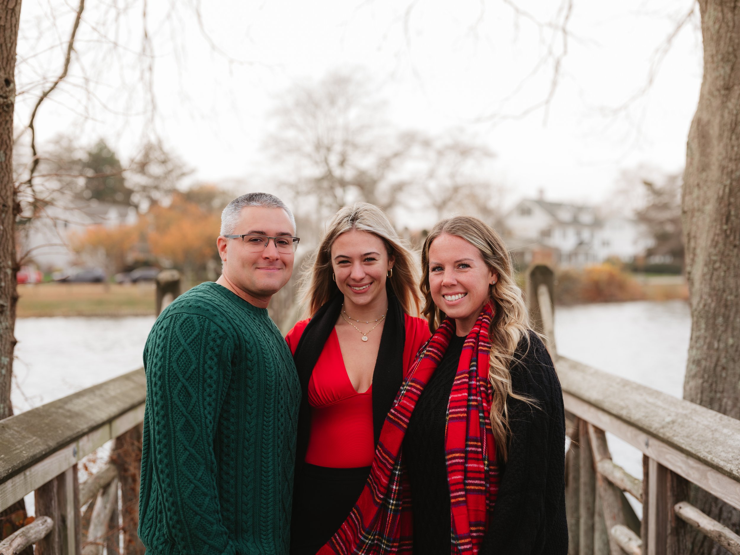Three people standing on a wooden bridge near a river, with trees and houses in the background, during fall.