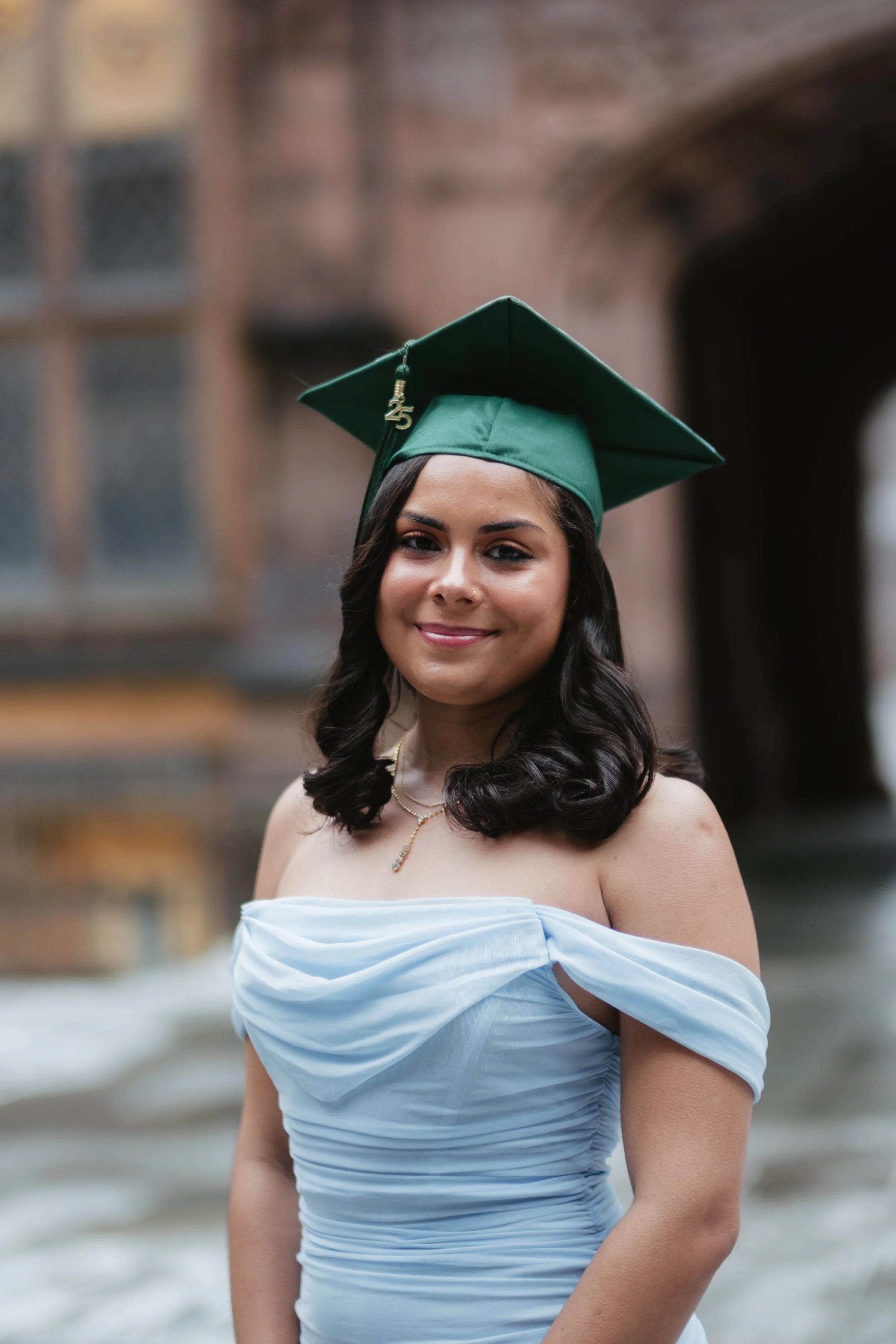 Young woman in off-shoulder blue dress wearing a green graduation cap with the number 25 charm, smiling outdoors with a historic building in the background.