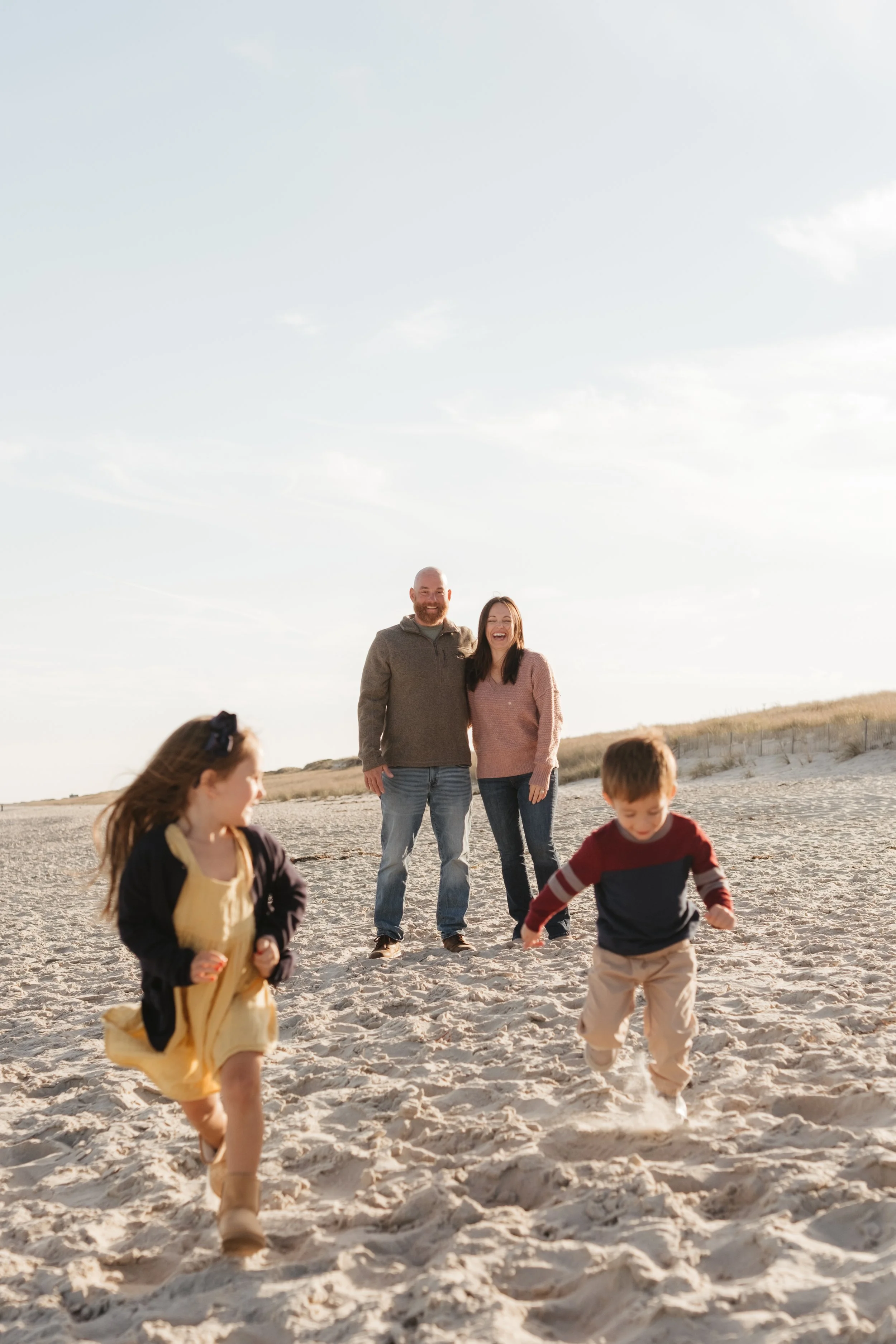 A family of four at the beach, with two children running in the sand and adults watching and smiling, under a clear sky.