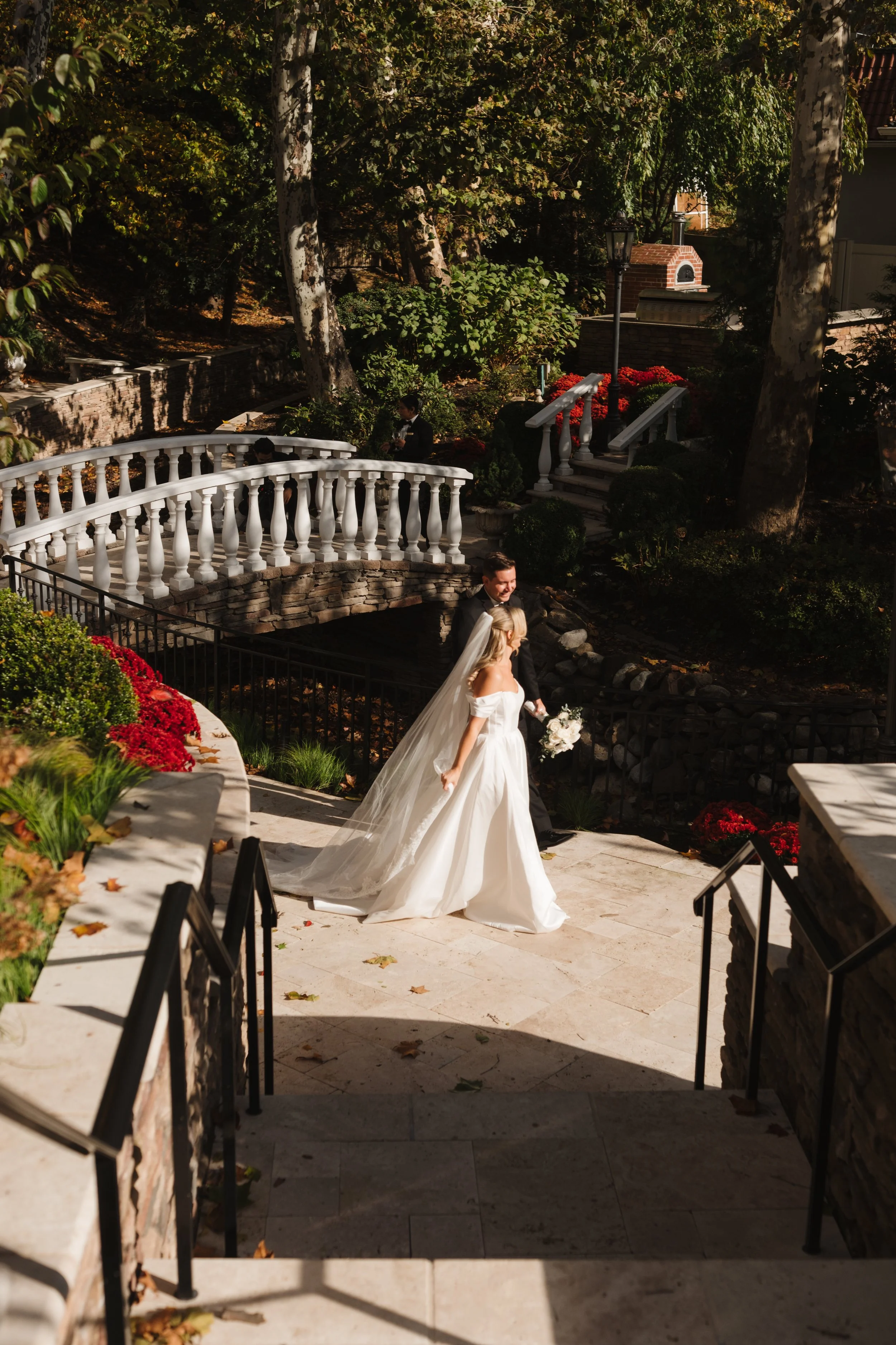 A bride and groom walk together down outdoor steps in a garden setting during a wedding ceremony, surrounded by greenery and colorful flowers.