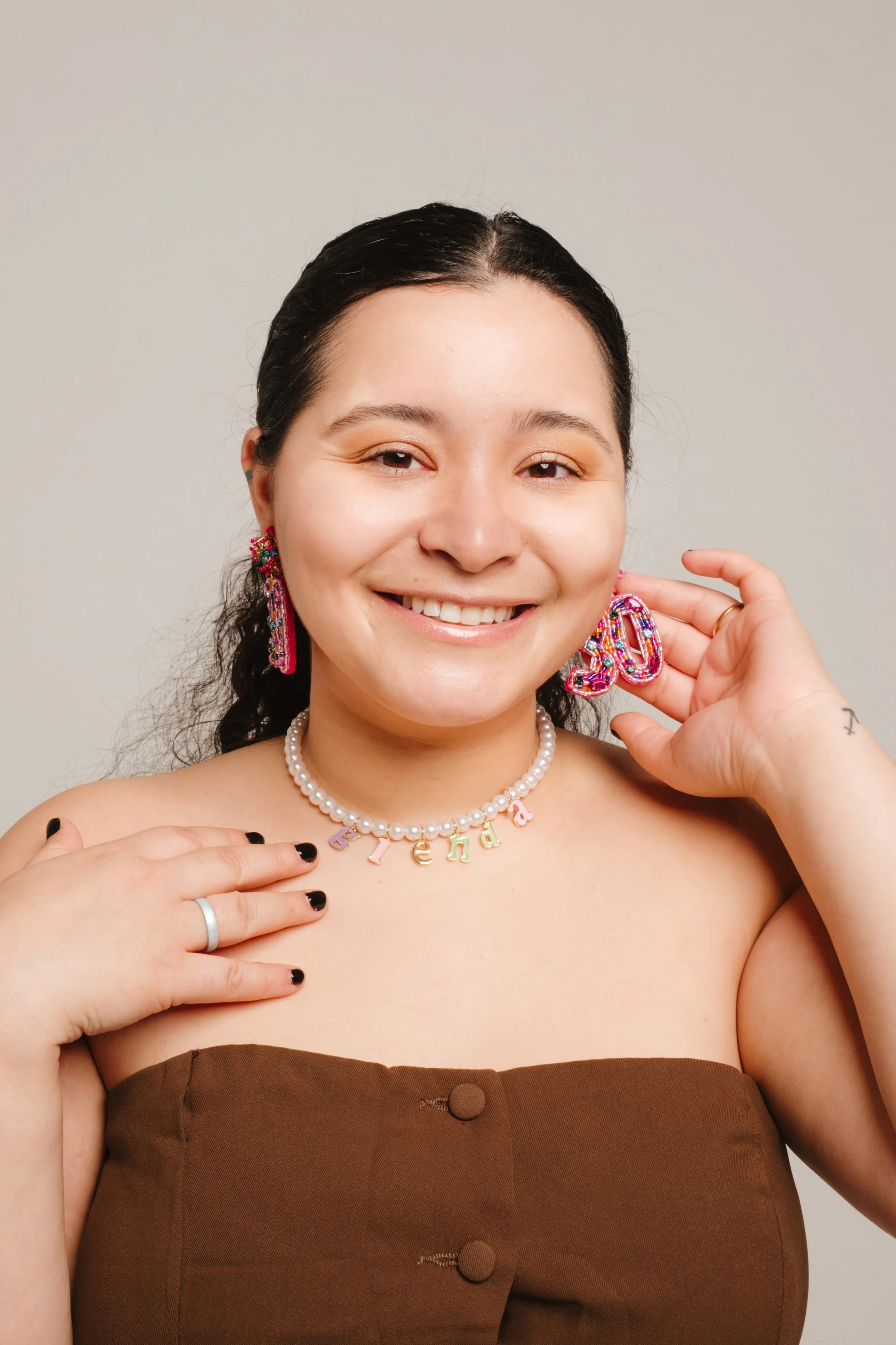 A young woman with dark hair in a ponytail smiling, wearing colorful jewelry including earrings and a pearl necklace with multicolored letter charms spelling 'Stelda', and a brown strapless top.