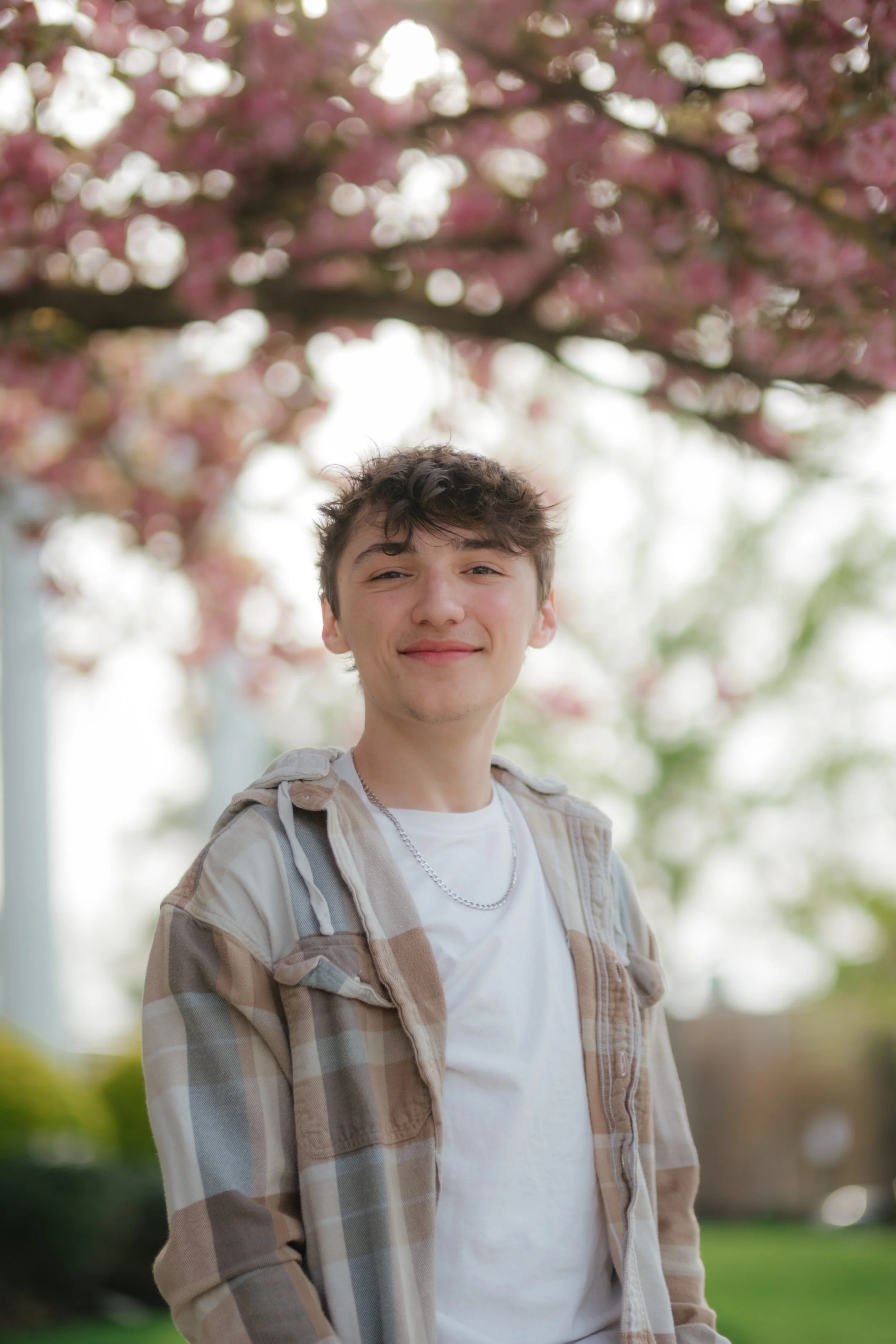 A young man smiling outdoors under a flowering tree with pink blossoms.