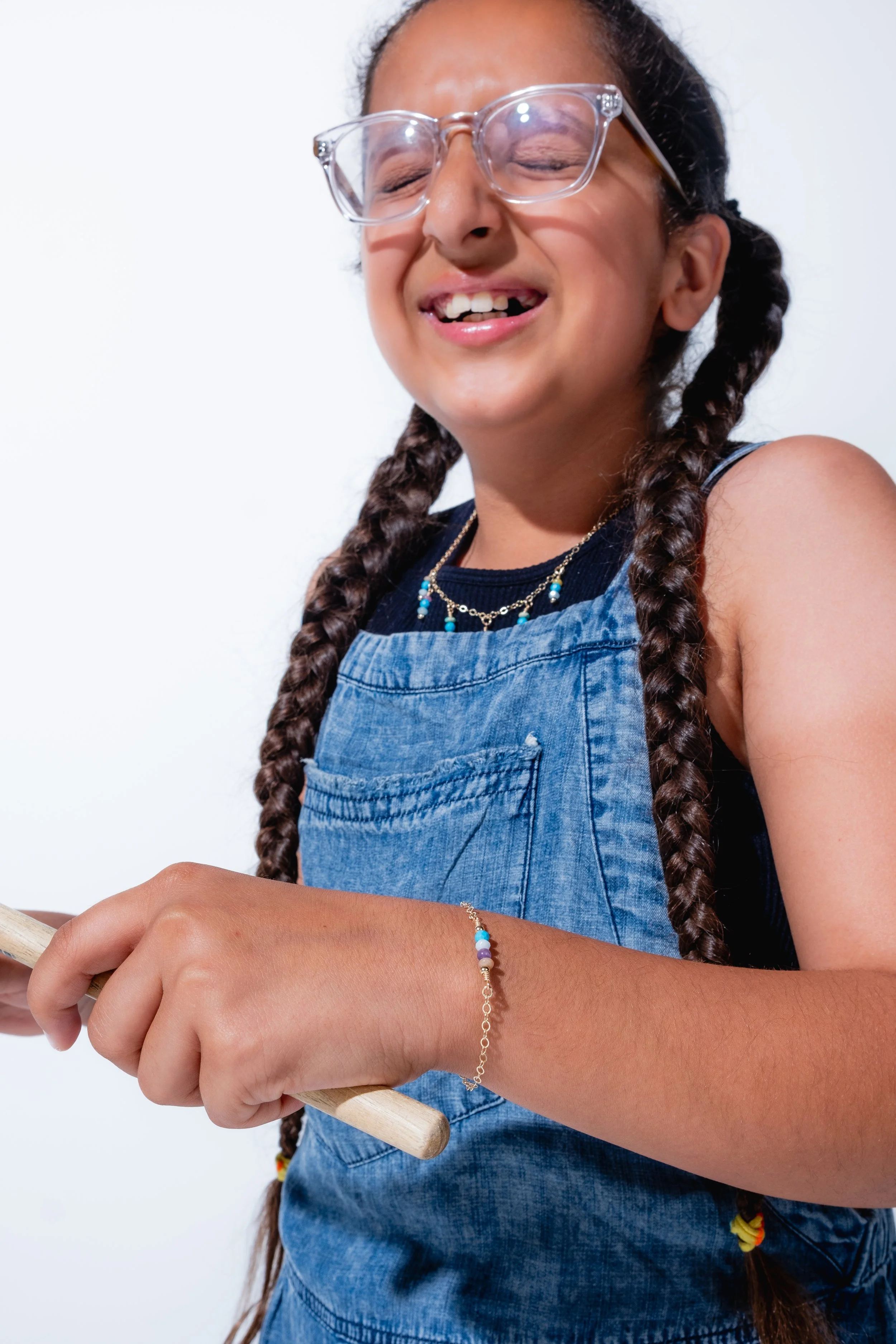 A happy girl with braided hair, wearing glasses, jewelry, and a denim overall, smiling and holding a wooden stick.