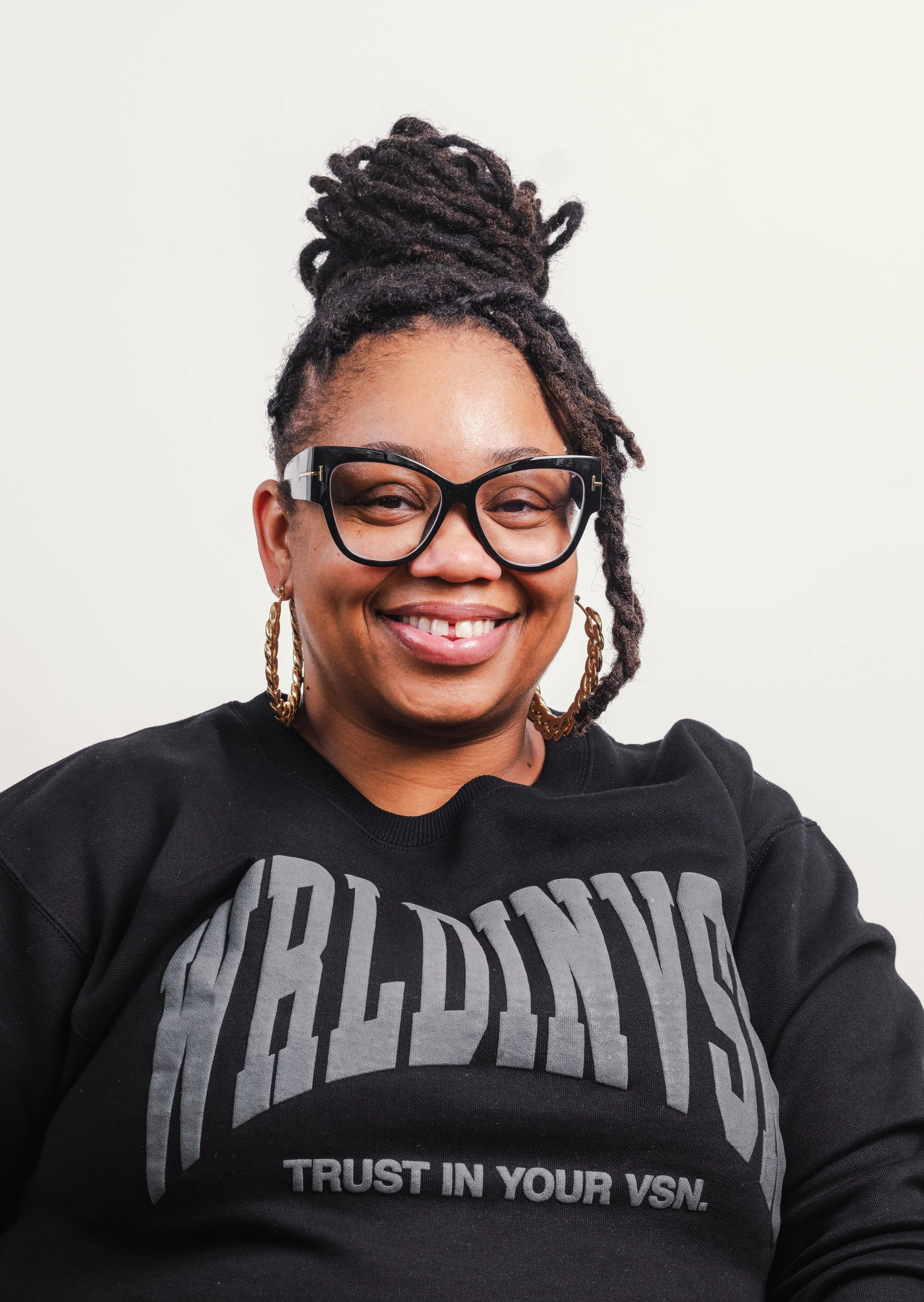 A smiling woman with glasses and large hoop earrings, wearing a black sweatshirt with bold gray text that reads 'VSL' and smaller text 'Trust in Your VSN,' against a plain white background.
