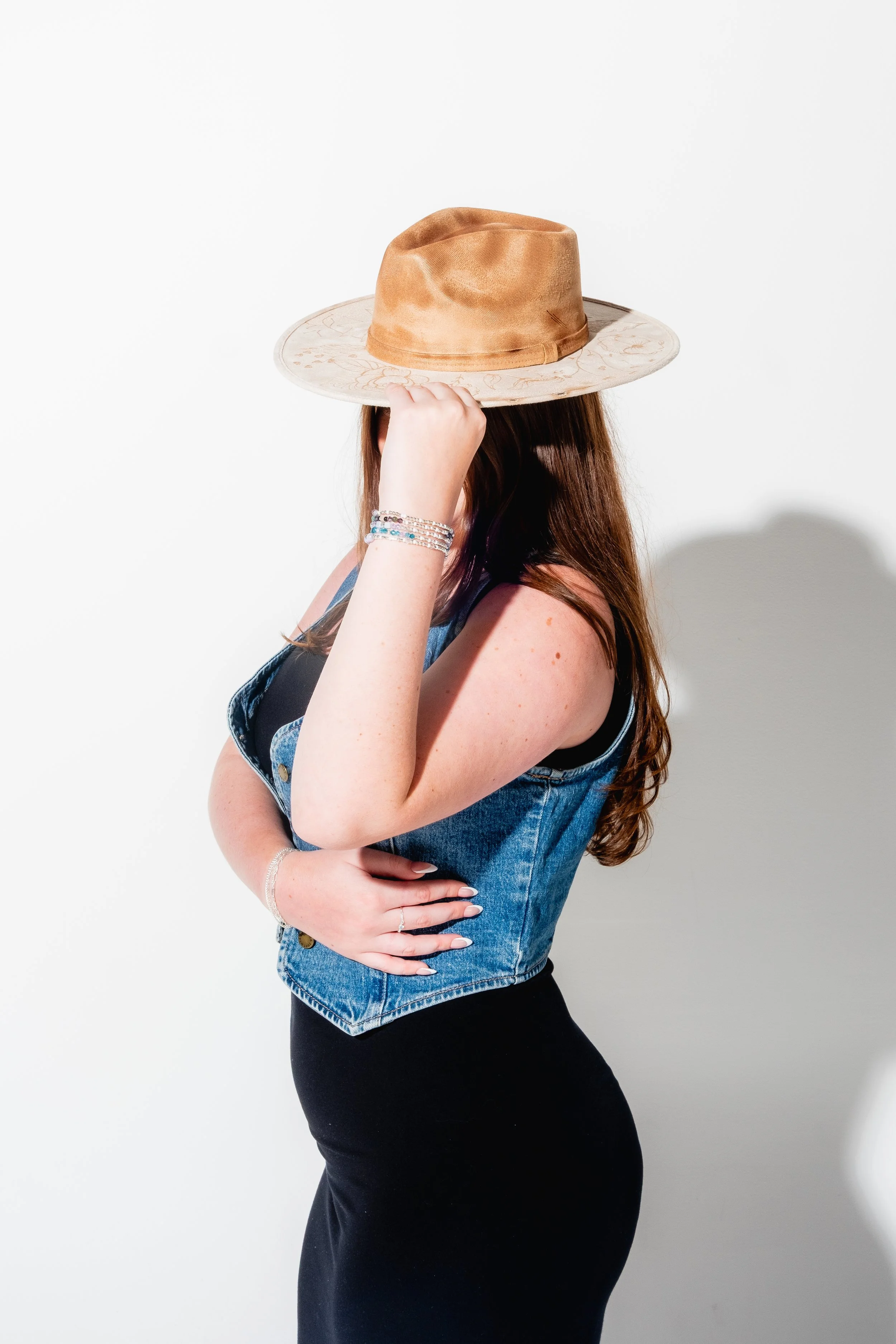 A woman with long red hair wearing a denim vest, black dress, bracelets, and rings, holding a tan wide-brimmed hat on her head, against a white background.
