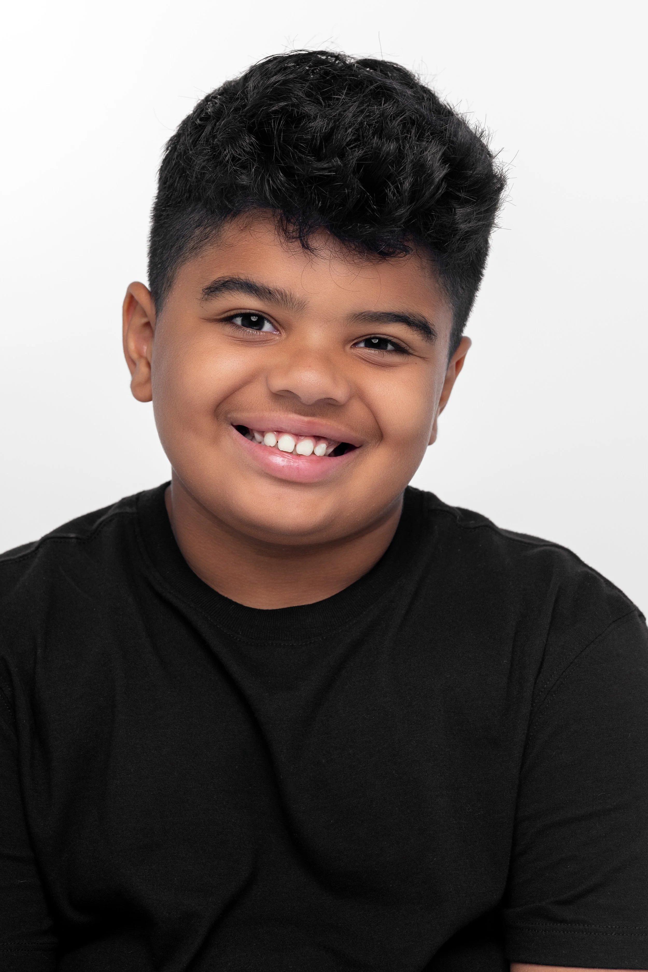 Smiling young boy with short dark curly hair, wearing a black shirt, against a plain white background.