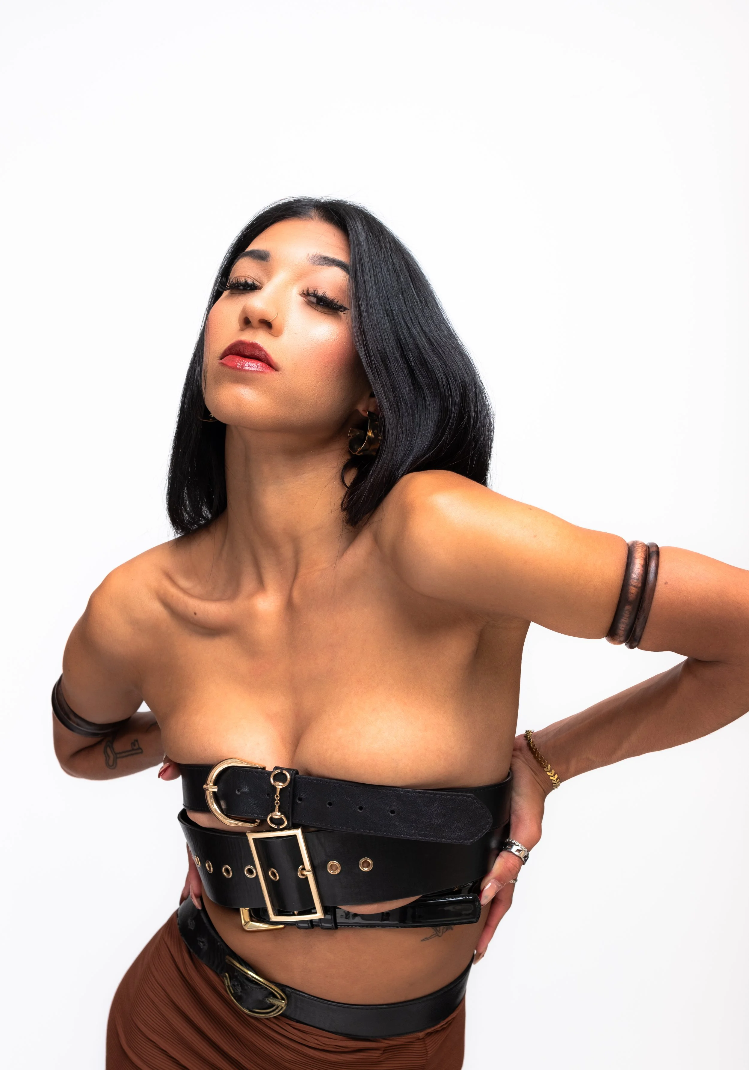 A woman with black hair, wearing a black belt, and brown pants, posing against a white background.