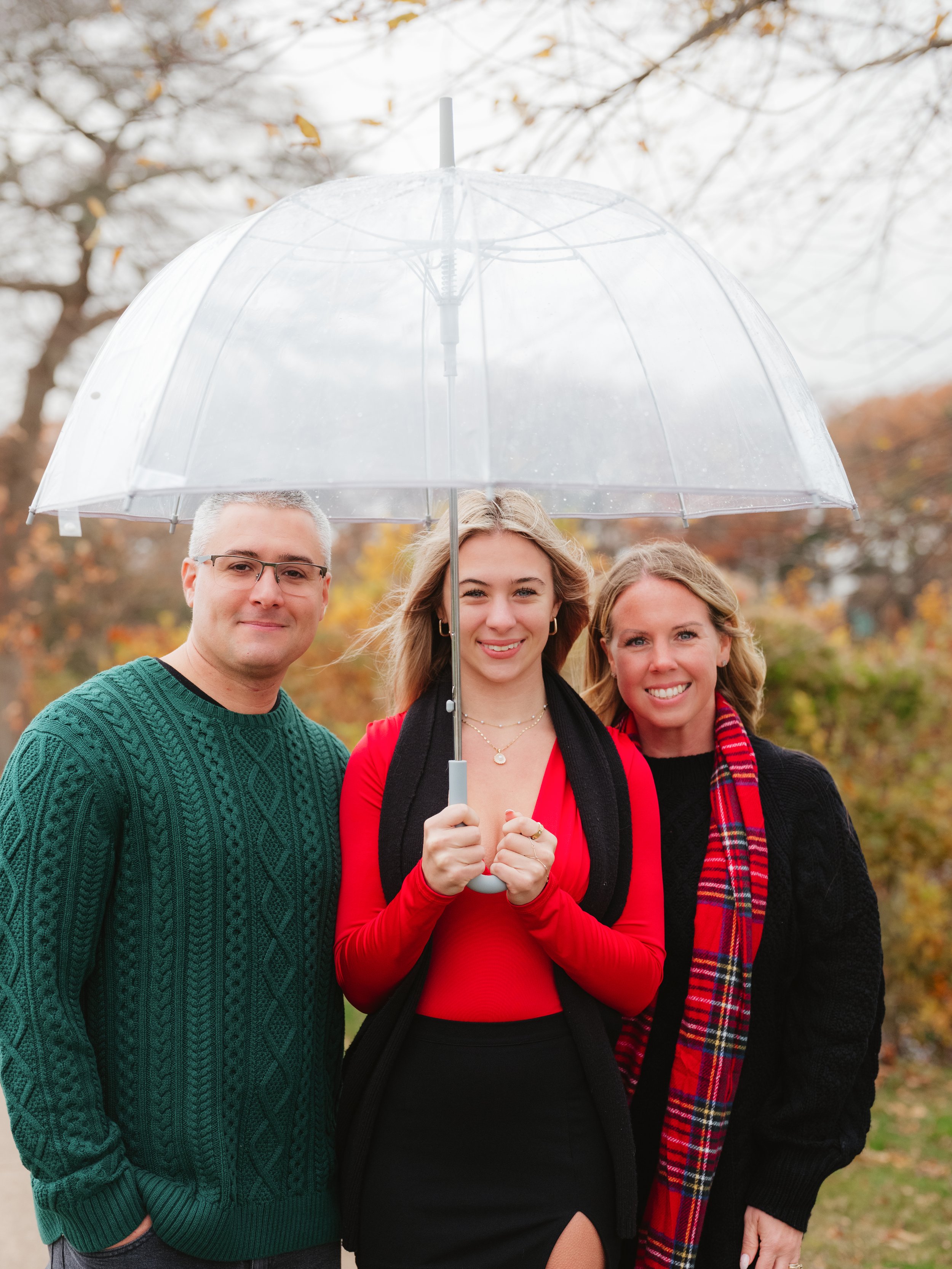 Three people standing outdoors, holding a clear umbrella, with fall foliage in the background. The woman in the center is wearing a red top and black skirt, smiling, while the man on the left in a green sweater and the woman on the right in a black s