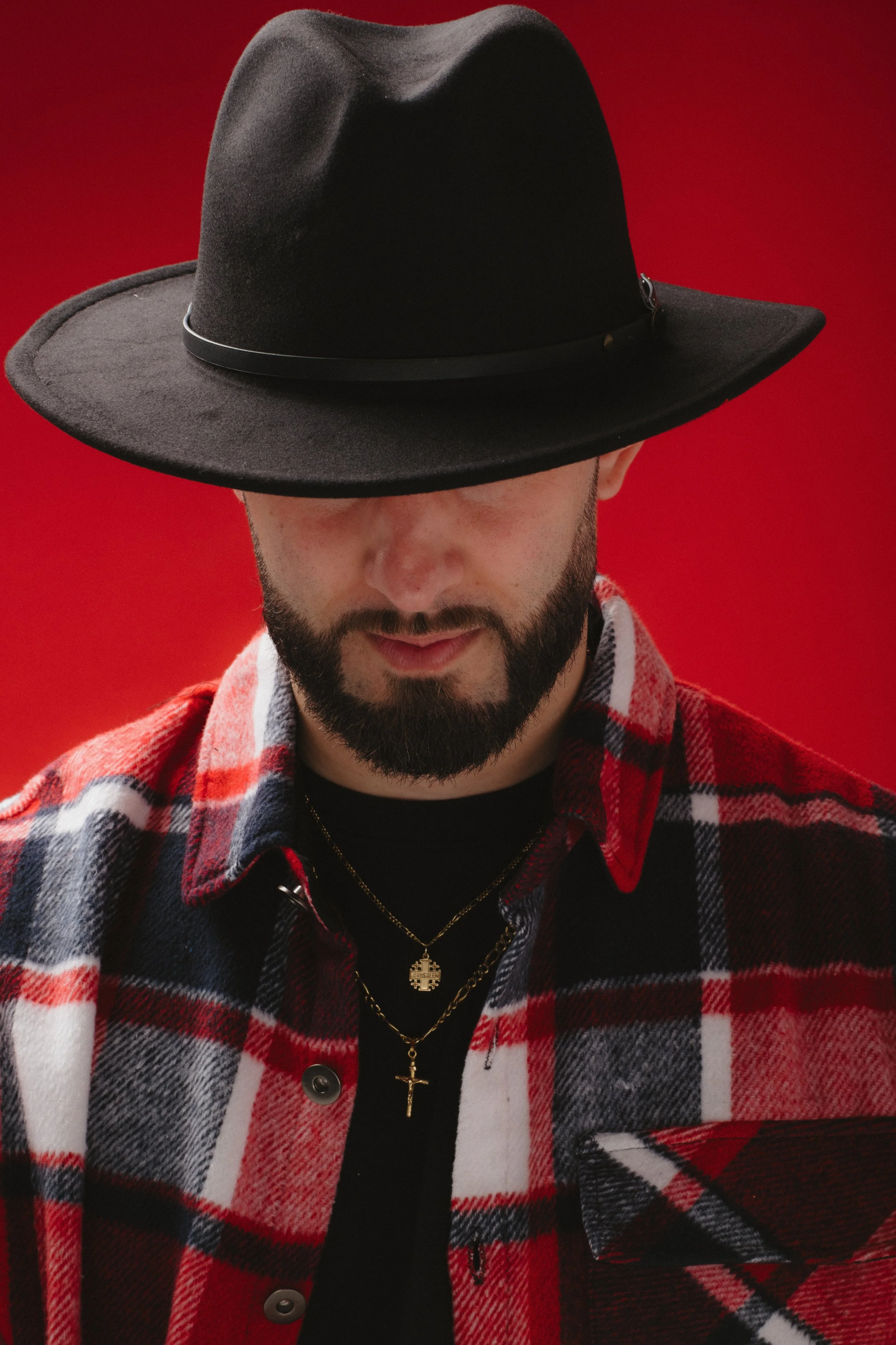 A man wearing a wide-brimmed black hat, a red and black plaid shirt, and layered gold necklaces with pendants, against a red background.