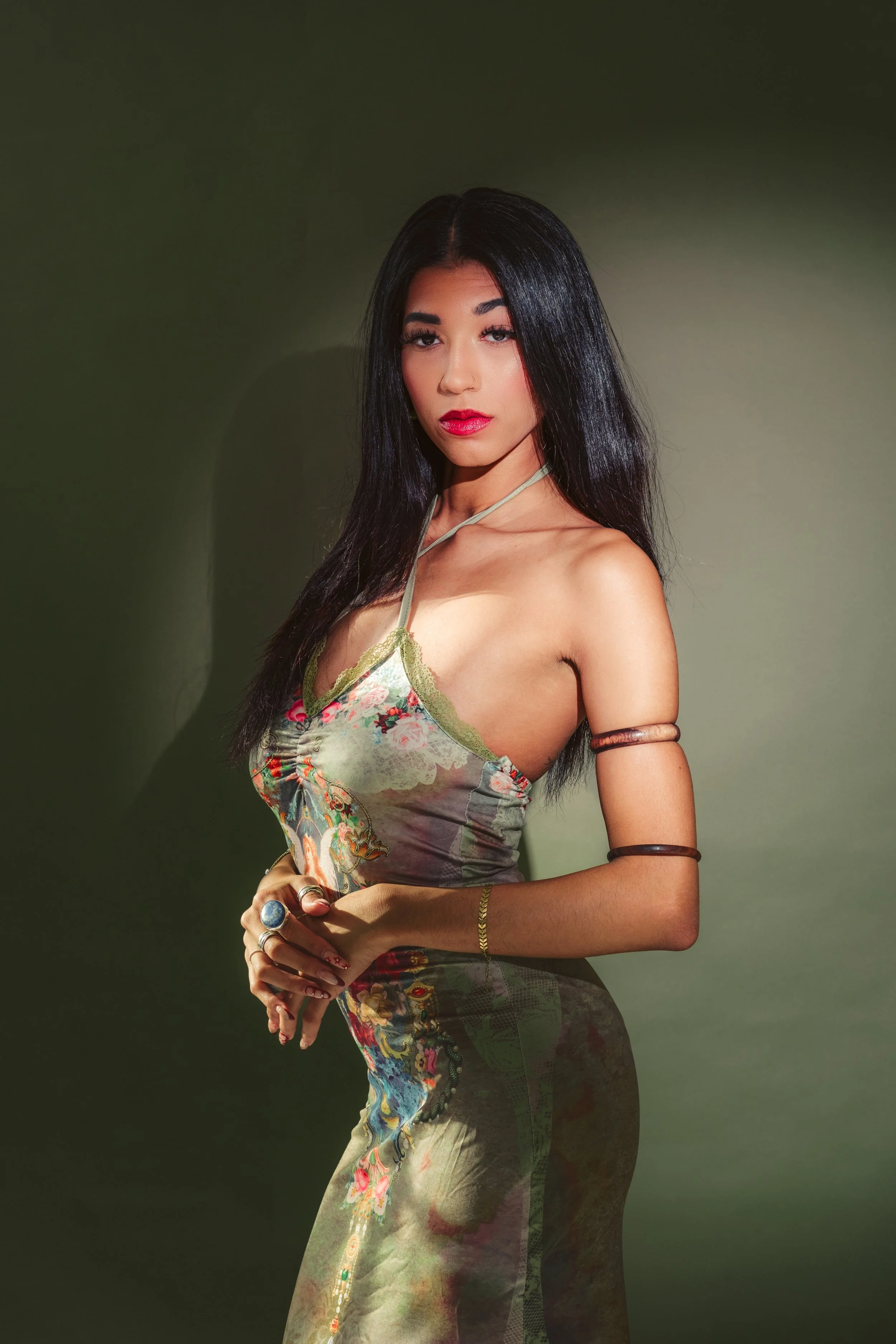 A woman with long black hair and red lipstick posing against a green background, wearing a colorful dress and jewelry.