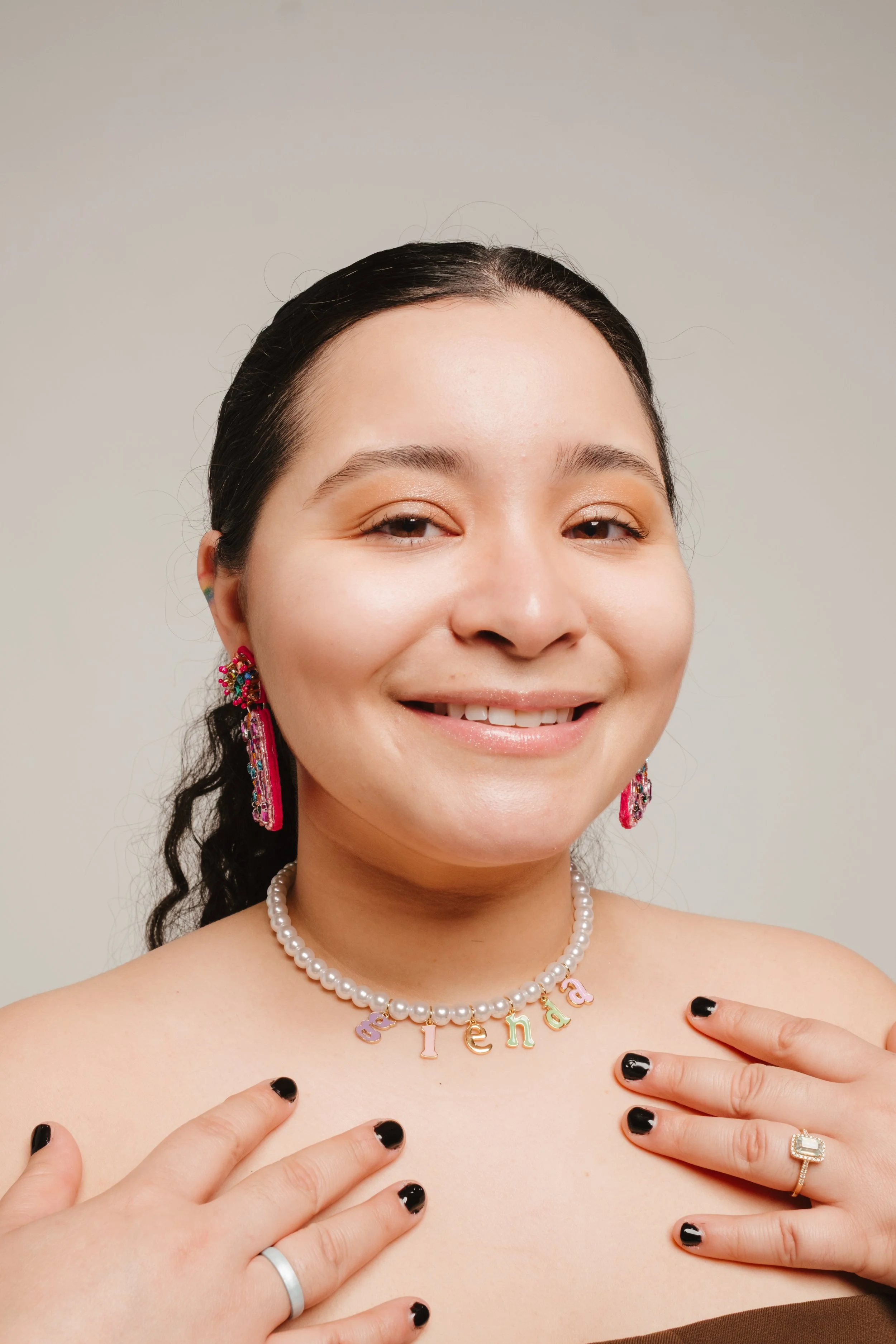A smiling woman with dark hair styled in a ponytail, wearing pink earrings, a pearl necklace with colorful letters spelling 'pende,' and rings on her fingers, holding her hands near her shoulders.