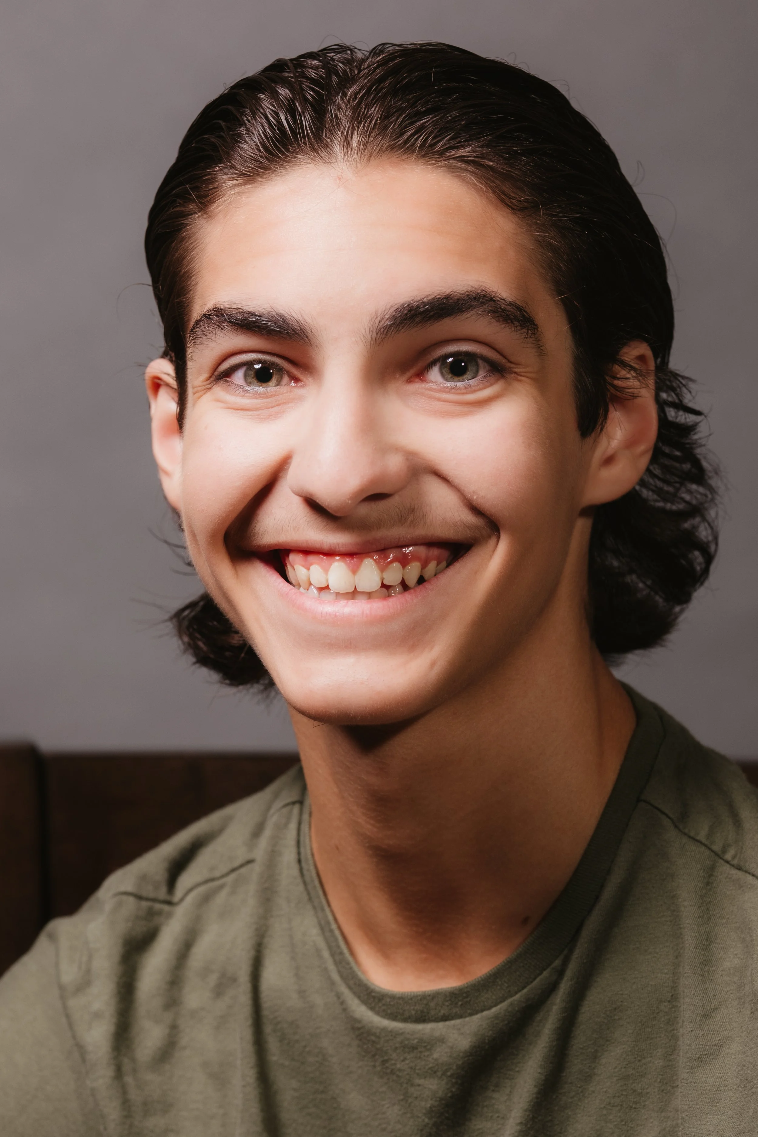 A young man with dark, slicked-back hair, bright eyes, and a big smile, wearing a green T-shirt, sitting against a neutral background.