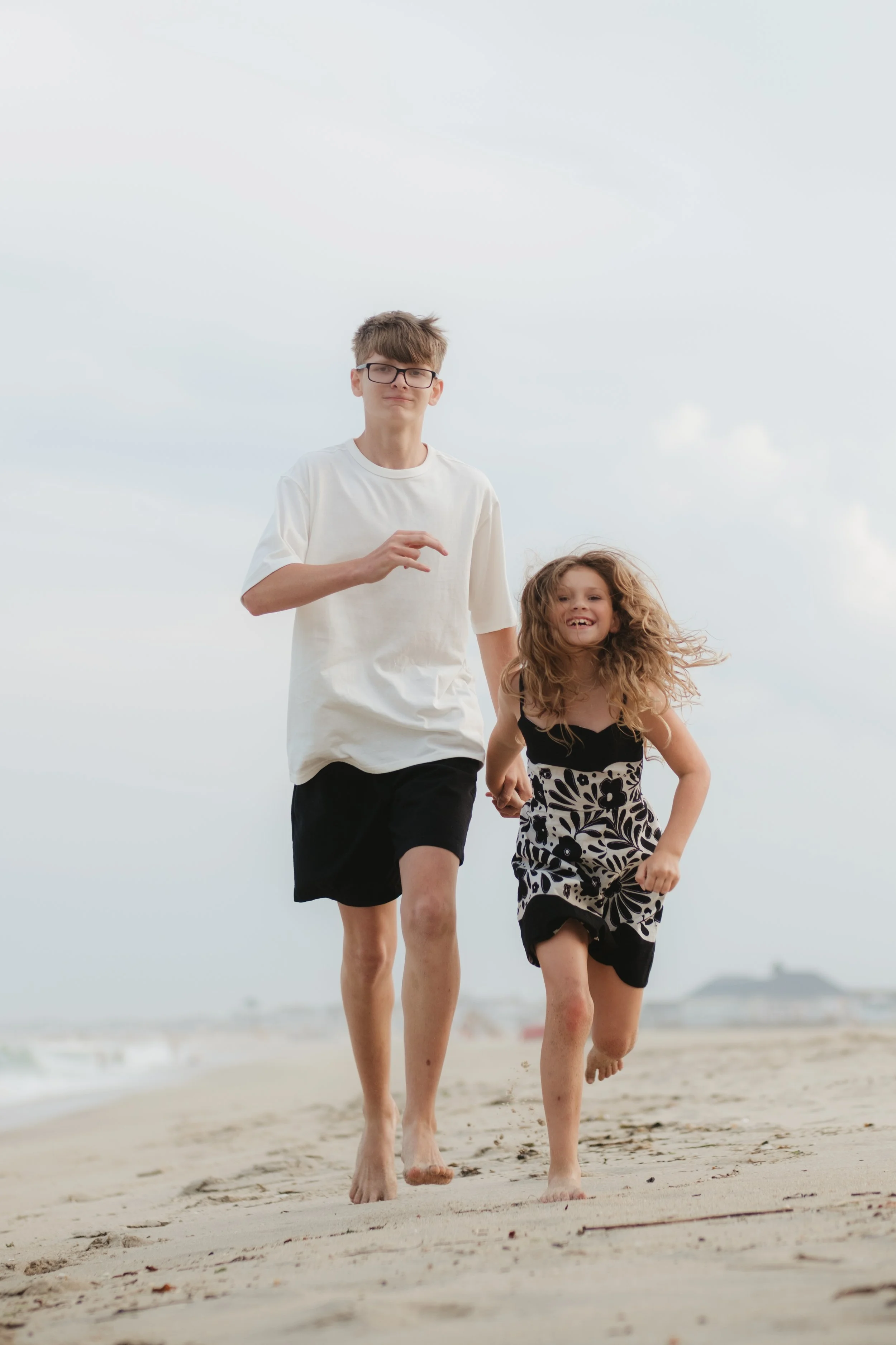 A young teenage boy and a young girl running barefoot on the beach, smiling, under a cloudy sky.