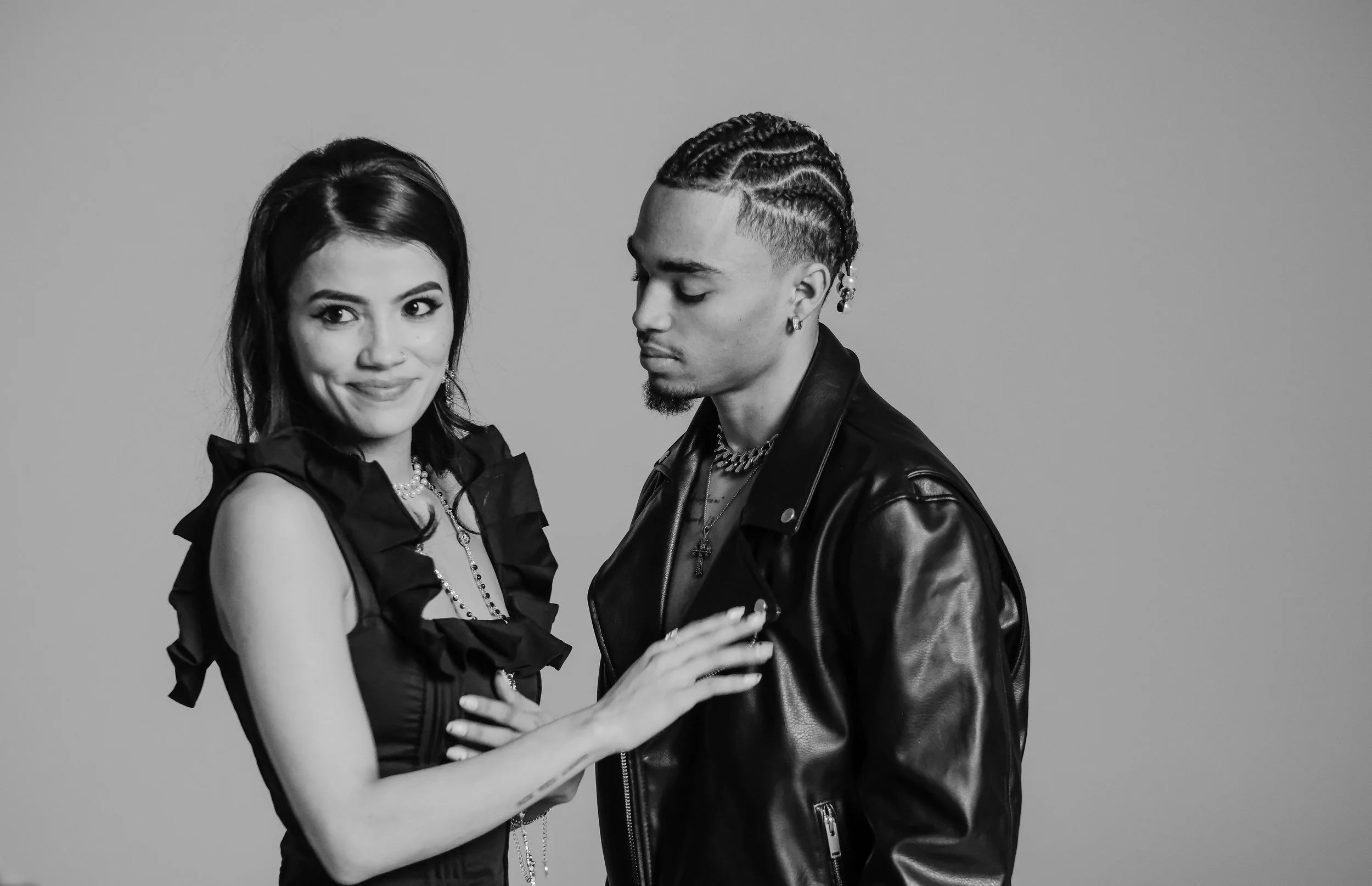 A black and white photo of a woman and man standing close together. The woman has dark hair, wears a sleeveless top with ruffles and jewelry, and is smiling at the camera. The man has cornrows, wears a leather jacket, and is looking down with a serio