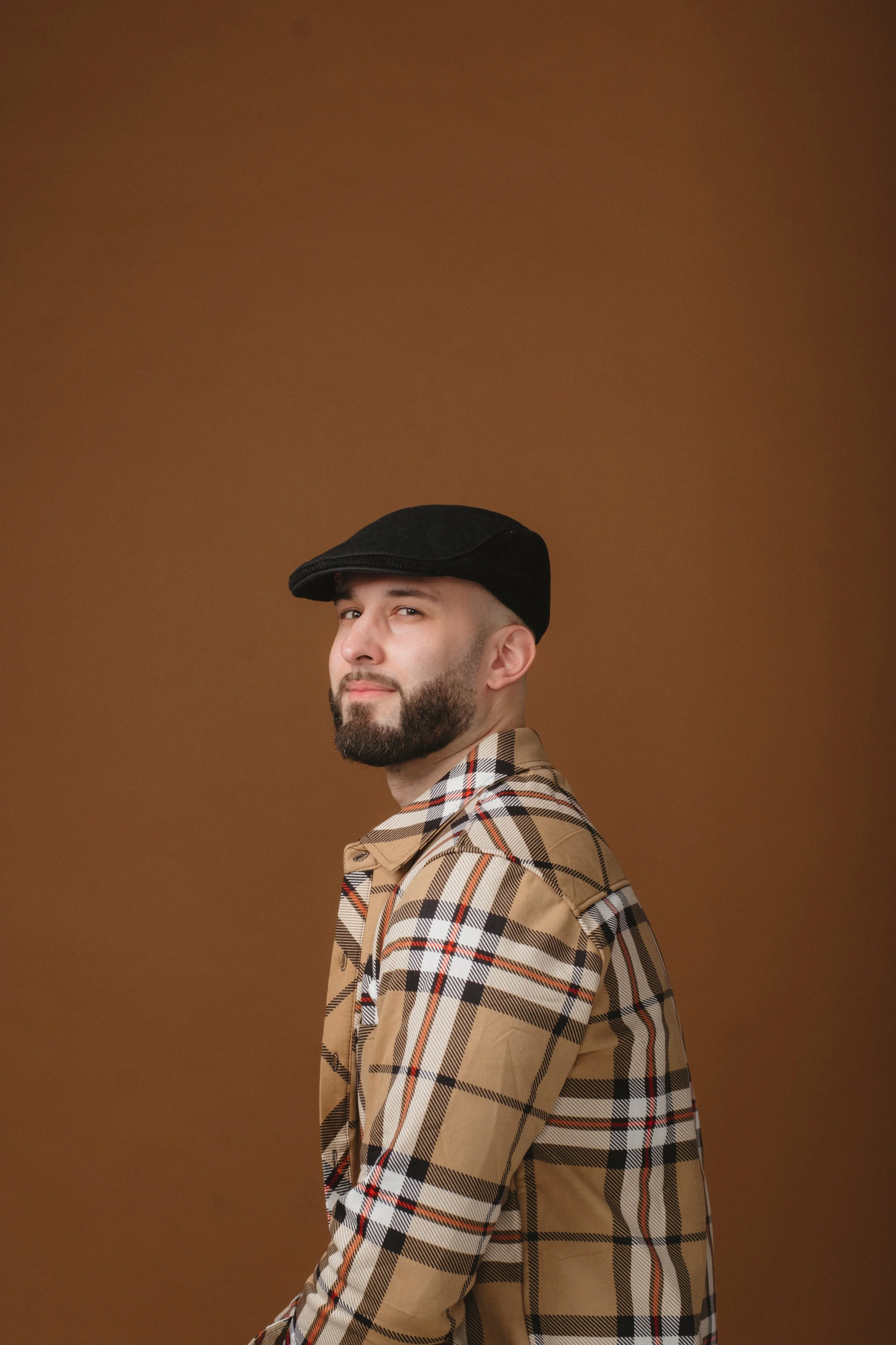 A man wearing a black cap and a beige plaid shirt, looking over his shoulder against a brown background.