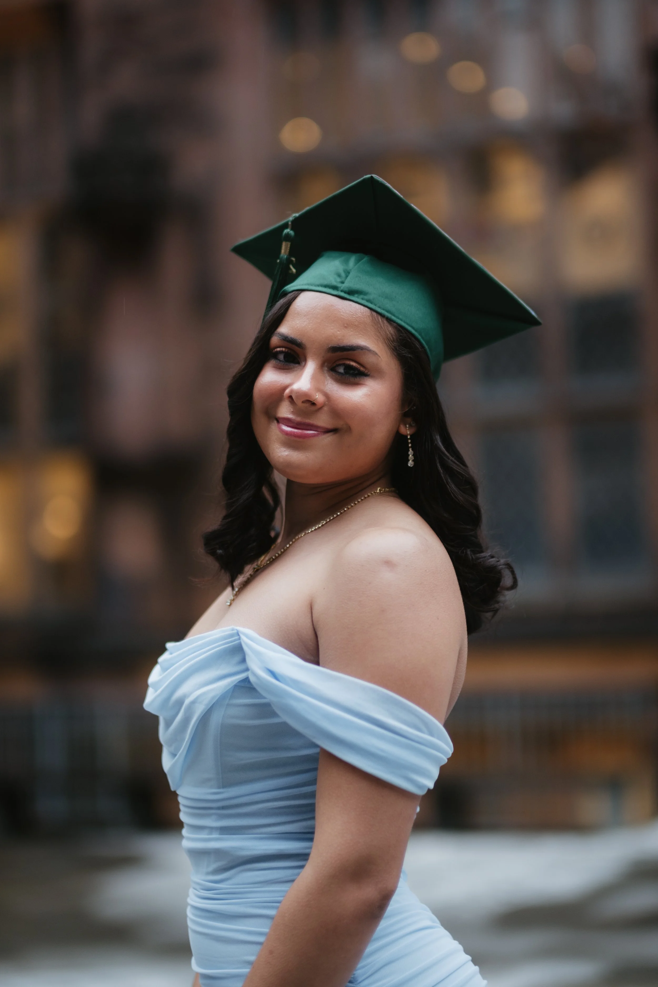 A woman wearing a green graduation cap and a light blue off-the-shoulder dress, smiling outdoors with a blurred background.