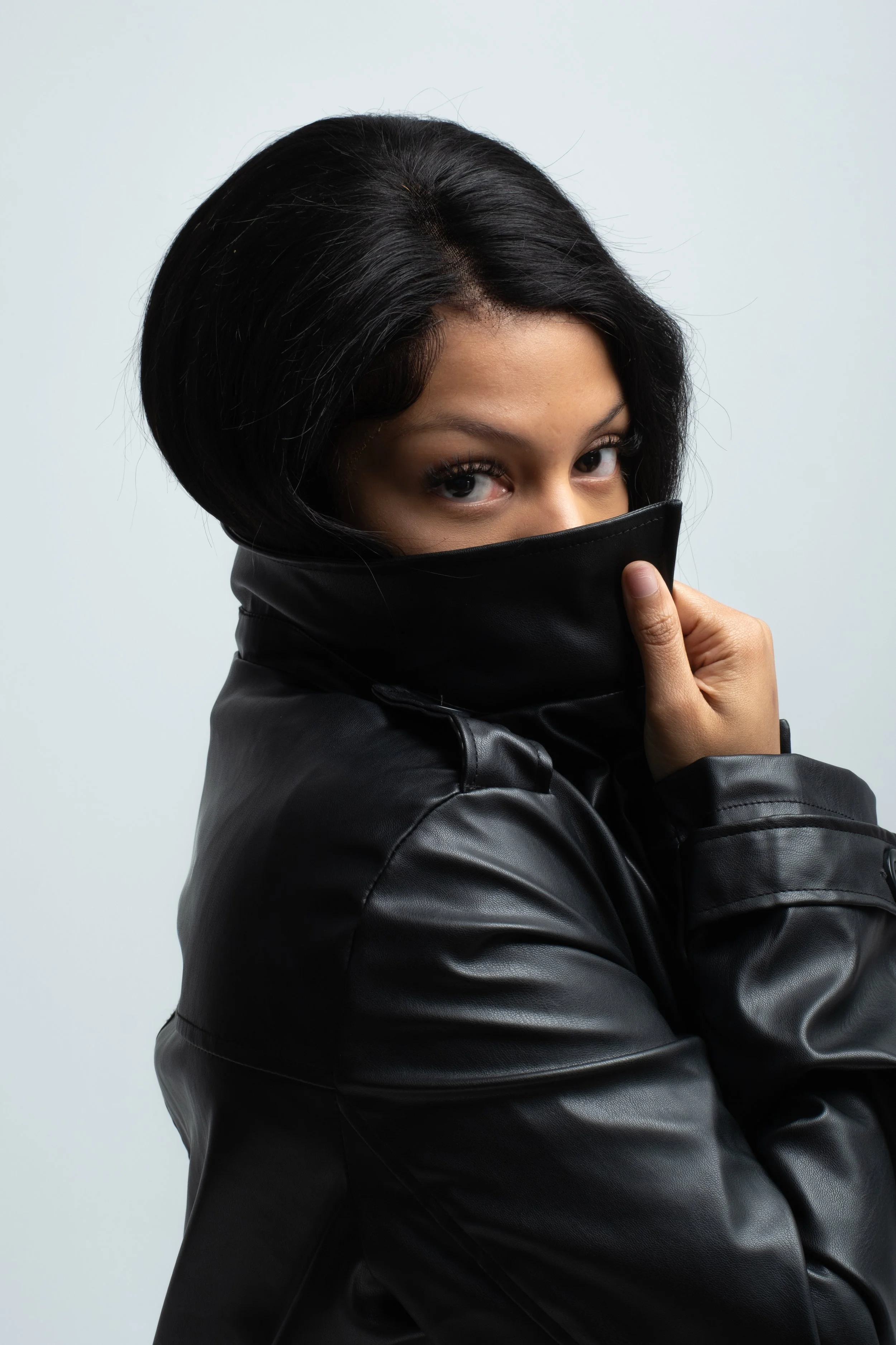 A woman with black hair wearing a black leather jacket and covering her mouth and nose with the collar, looking at the camera.