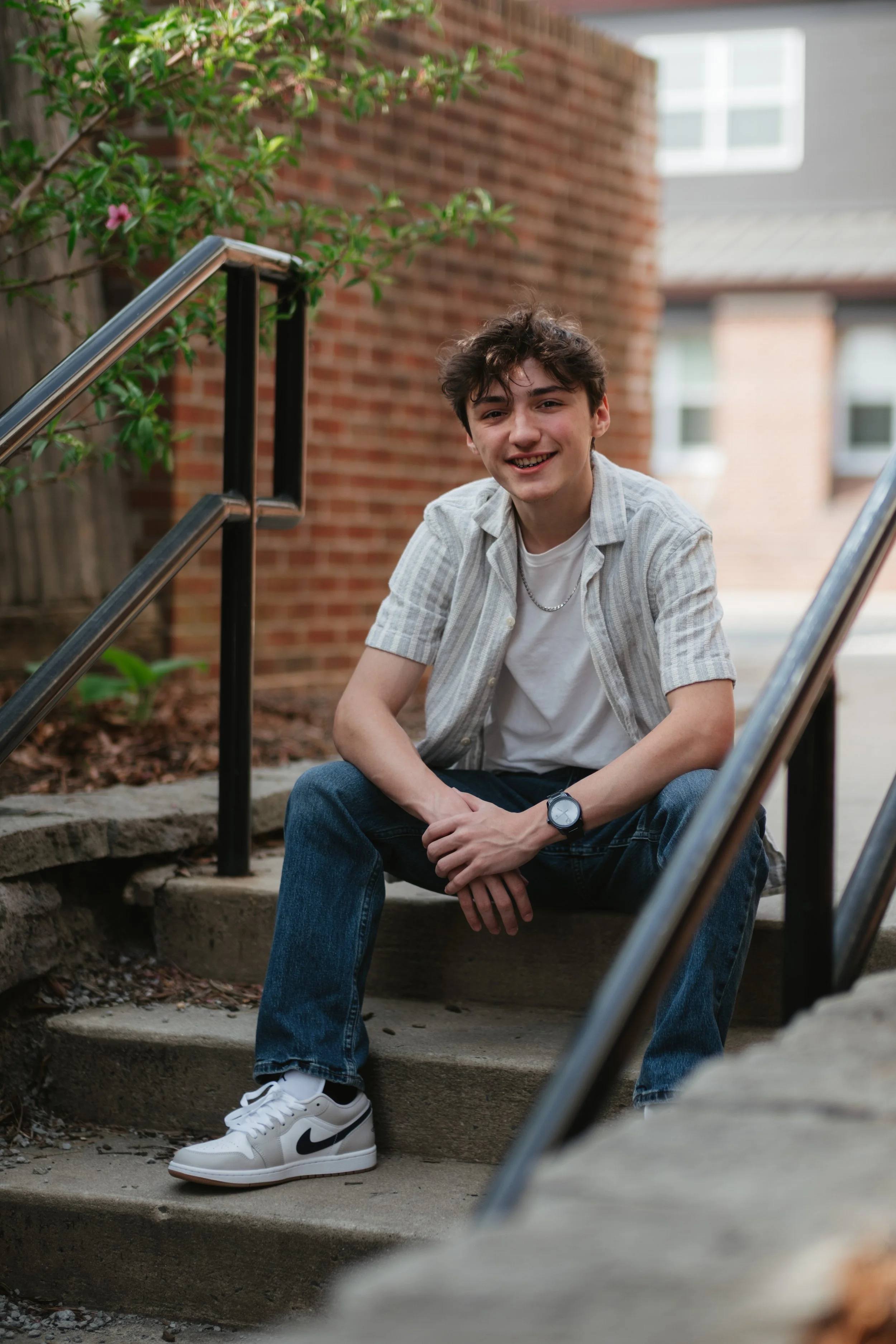 A young man sitting on outdoor concrete stairs next to a brick wall with greenery, wearing a casual short-sleeve button-up shirt, white T-shirt, jeans, white Nike sneakers, and a watch, smiling at the camera.