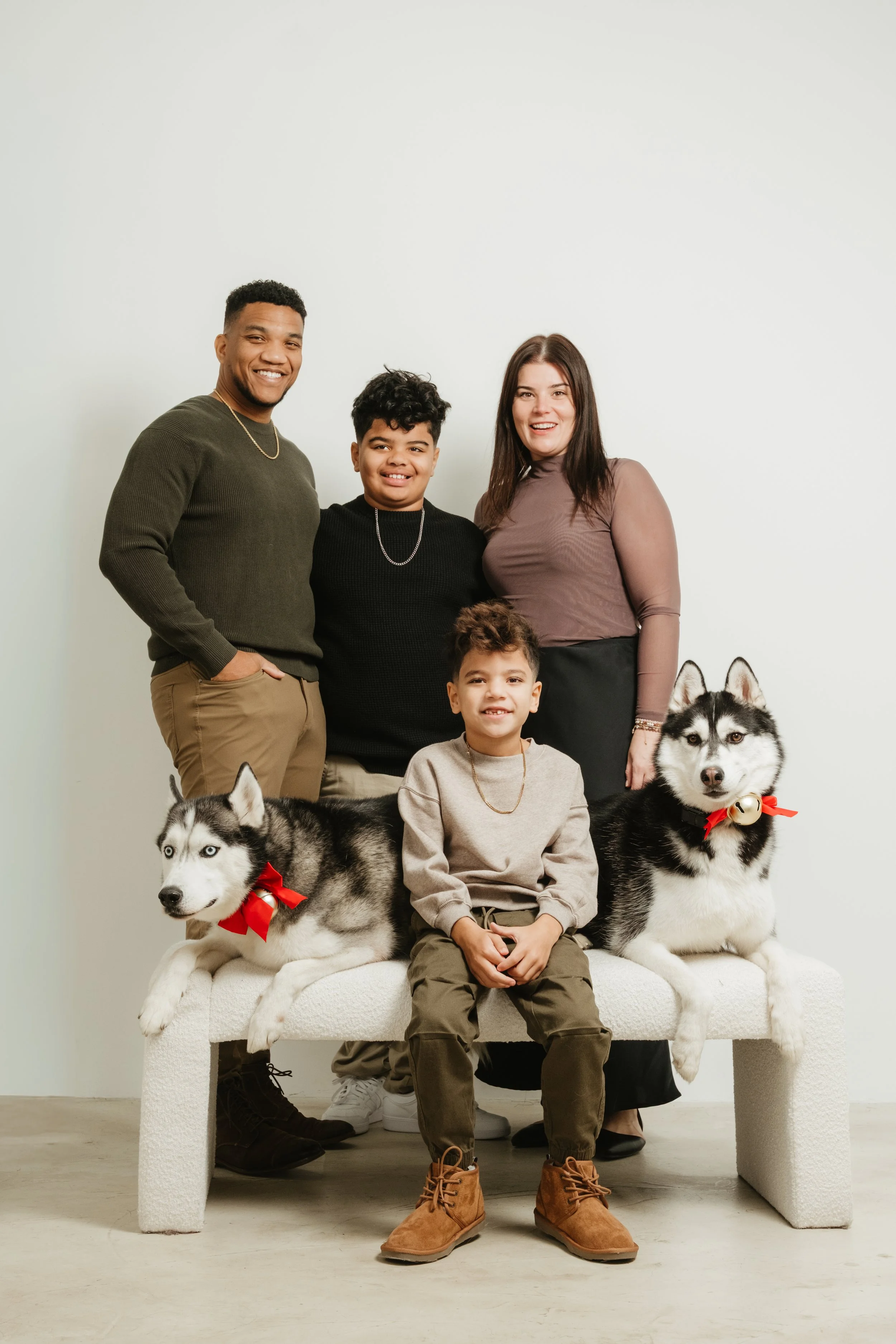 A family of four with two Siberian Huskies sitting on a white bench in front of a plain white background.